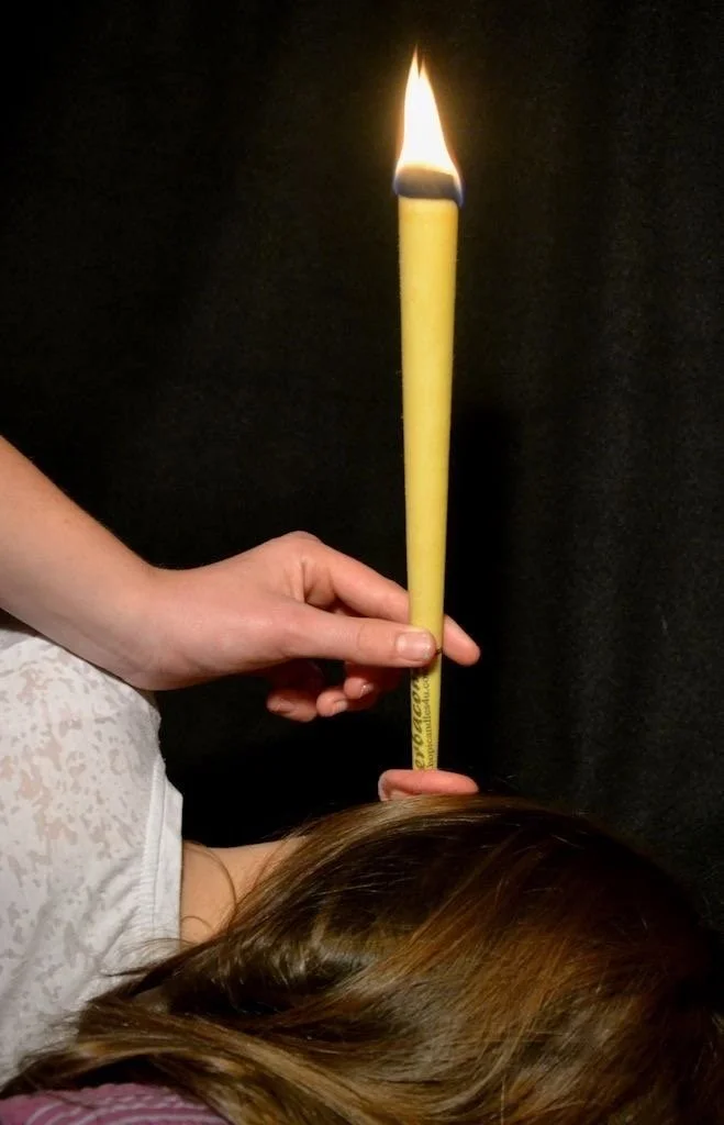 A person lying down holding a large yellow candle with a flame, ear candling, with hair partially visible and a dark background.