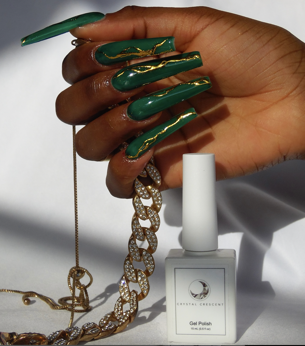 A person showing off long, green, gold-accented acrylic nails, holding a gold chain with rhinestones, with a white bottle of gel nail polish labeled 'Crystal Crescent' in front.