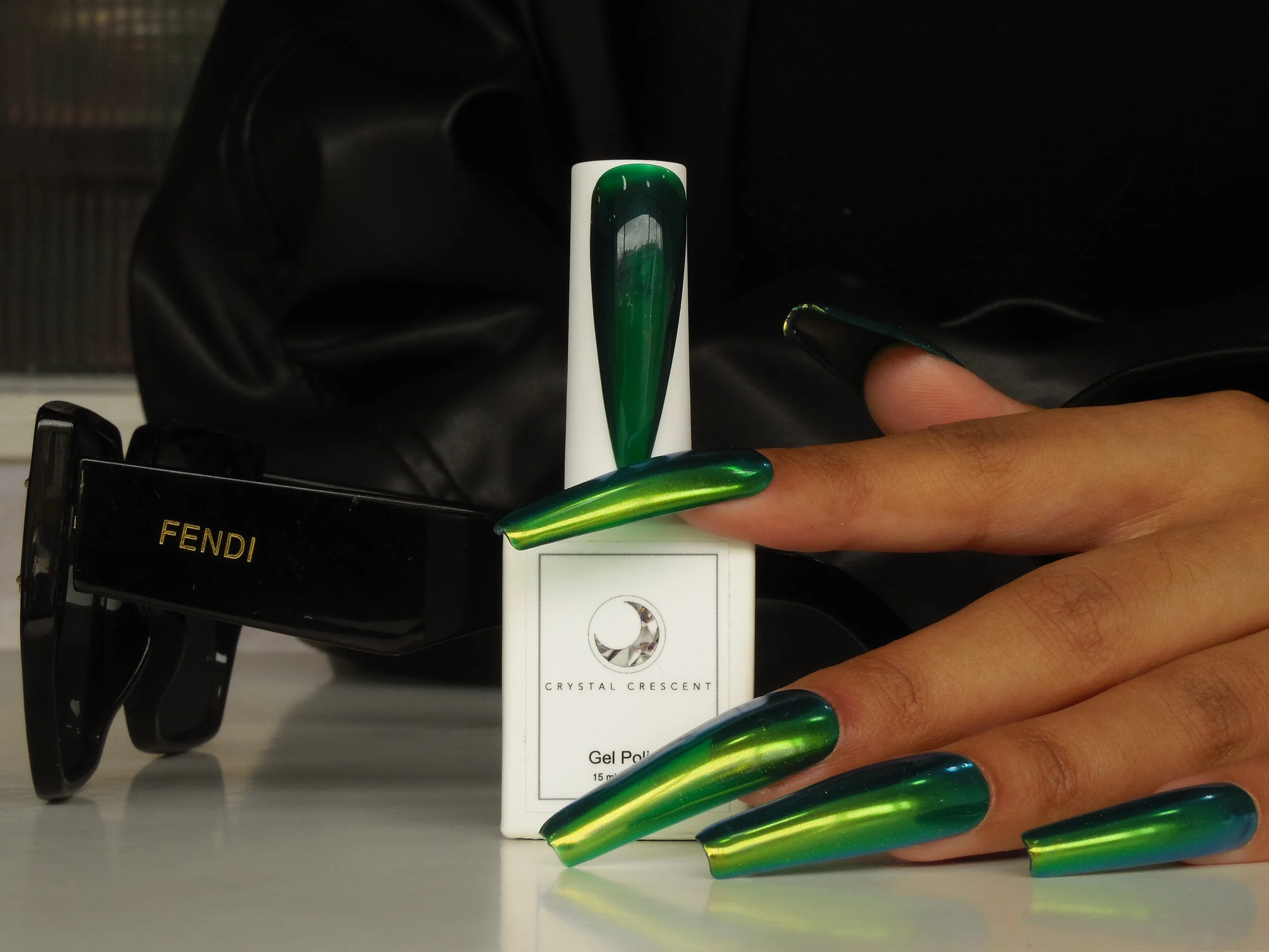 Hand with long, green and blue metallic nails, next to a small box of Crystal Crescent gel nail polish, and behind that, a black Fendi sunglasses case.