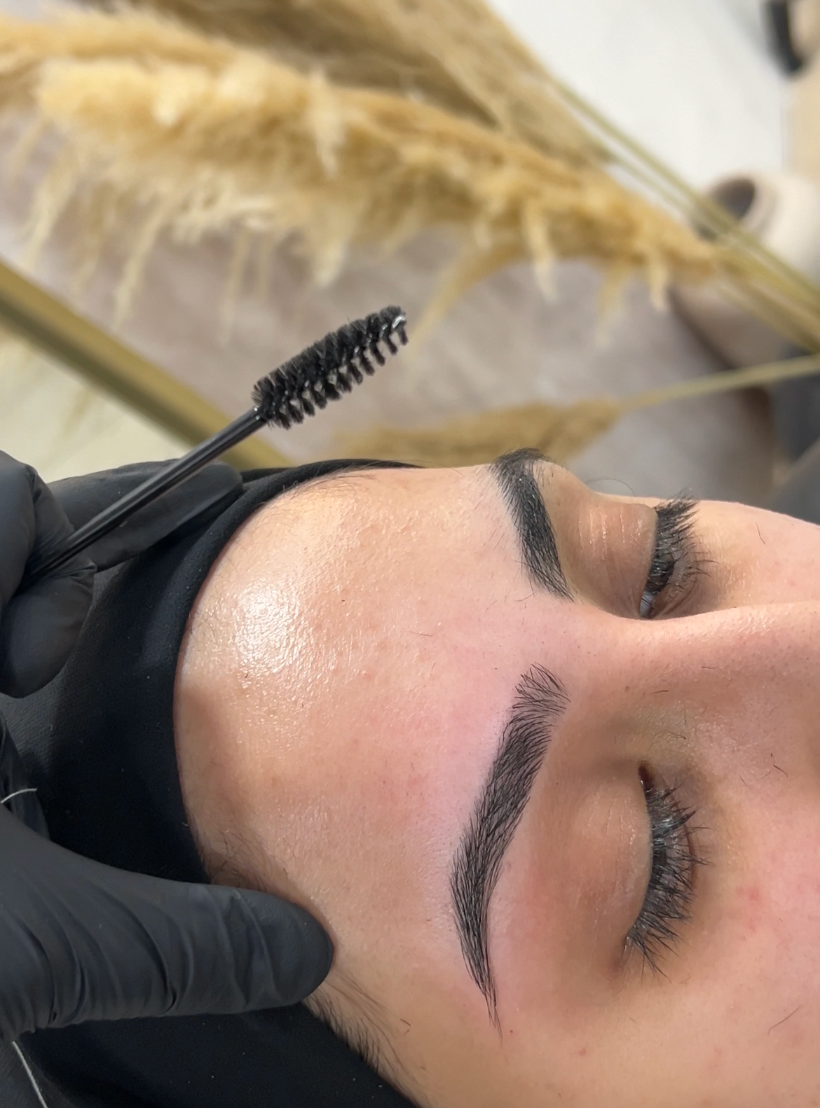 Close-up of a woman with freshly threaded eyebrows, lying down with her eyes closed, while a technician in black gloves applies touch-up with a brush.