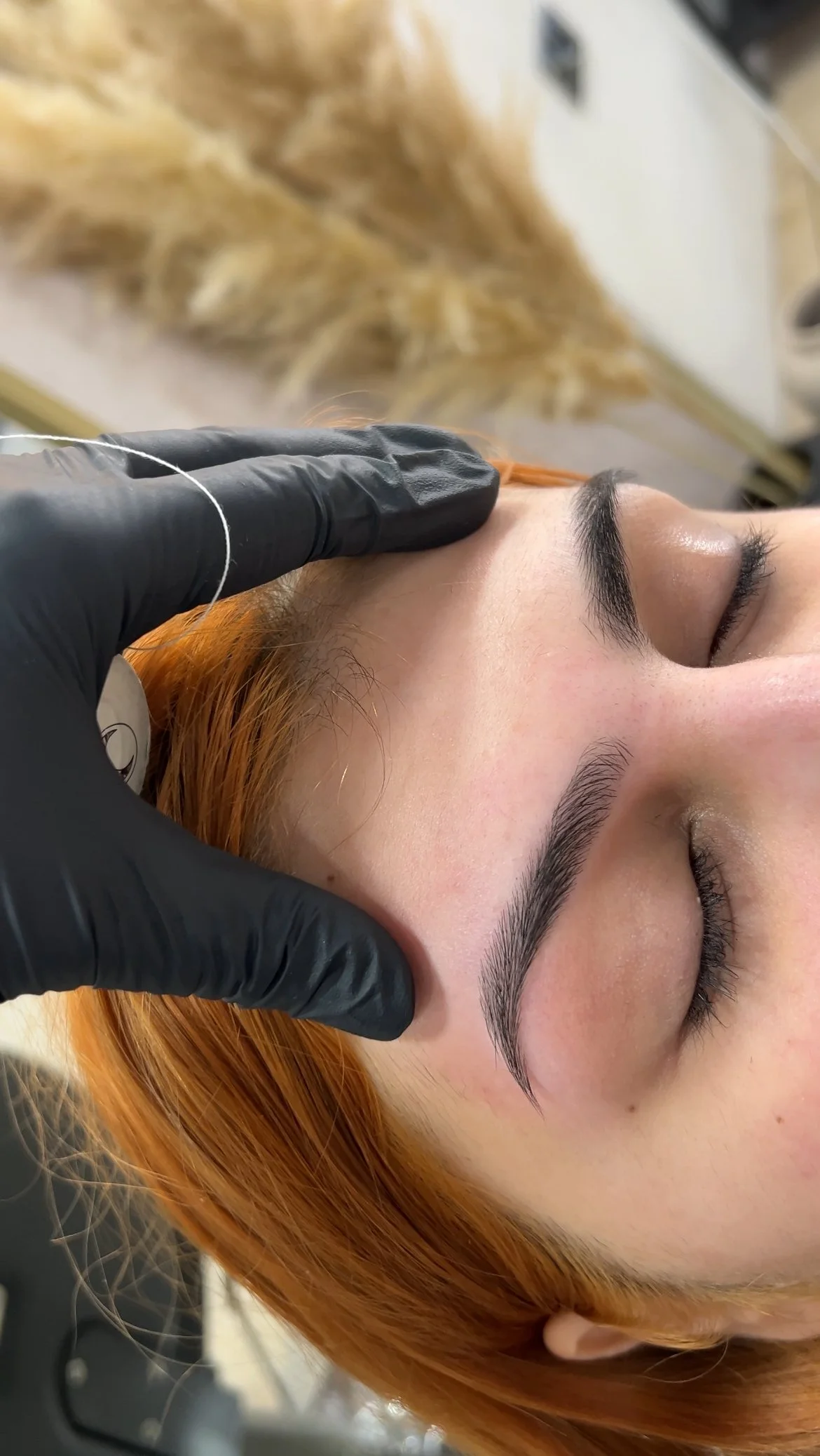 Close-up of a woman with freshly threaded eyebrows, lying down with eyes closed, while a technician in black gloves touches her forehead.