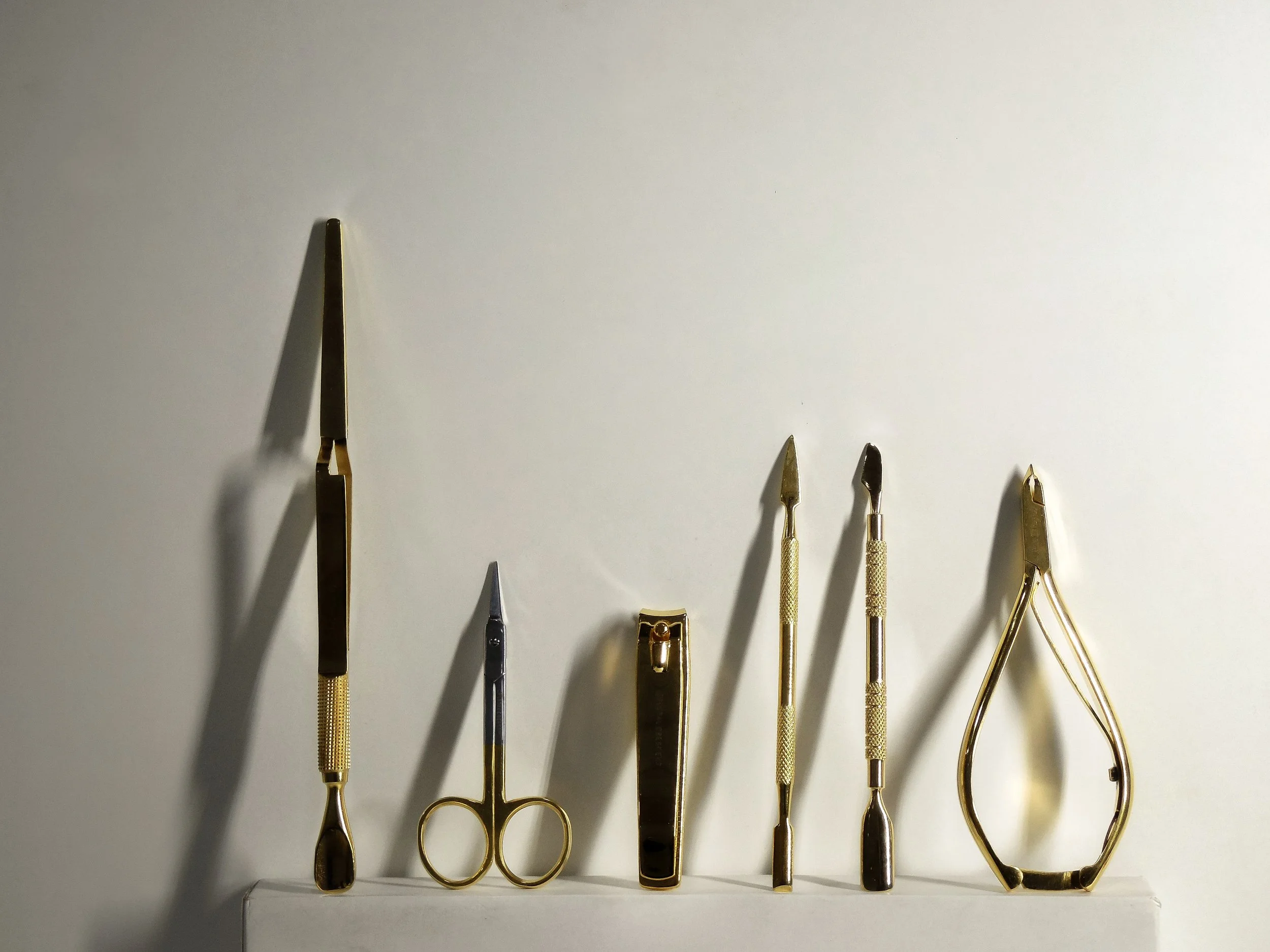 A collection of six nail care tools with gold handles, including a nail file, scissors, cuticle nippers, and other manicure instruments, arranged on a white surface against a white background.