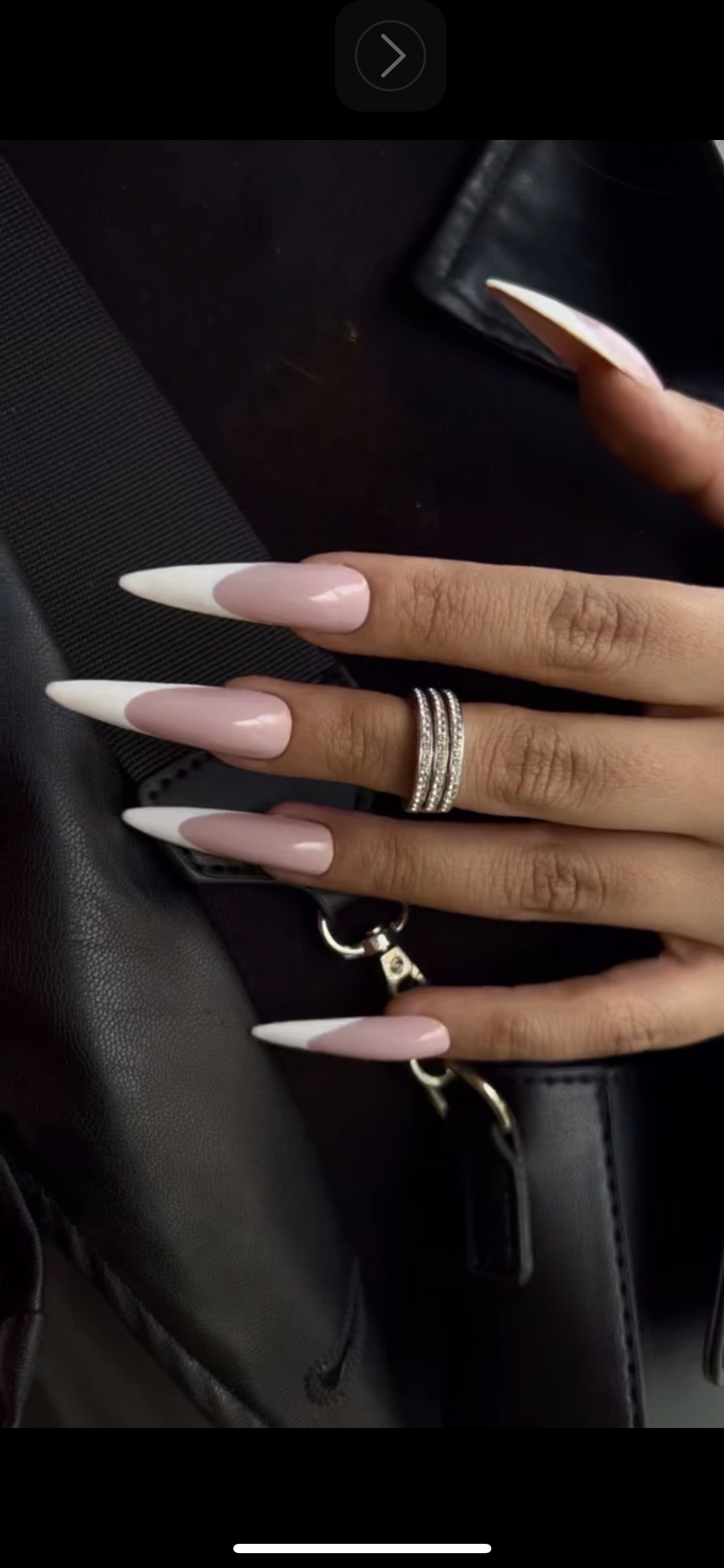 A person's hand with long acrylic nails painted in a light pink and white color, wearing multiple silver rings with small stones, holding a set of car keys, resting on a black leather surface with a wallet nearby.