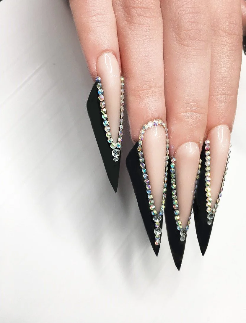 Close-up of long, pointed black and nude-colored acrylic nails decorated with rhinestones.
