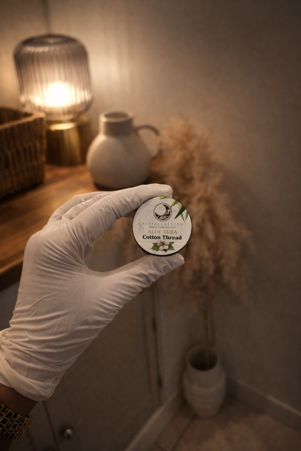 Hand in a glove holding a container of aloe vera cotton thread in a room with warm lighting, a wicker chair, a lamp, a vase, and decorative plants.