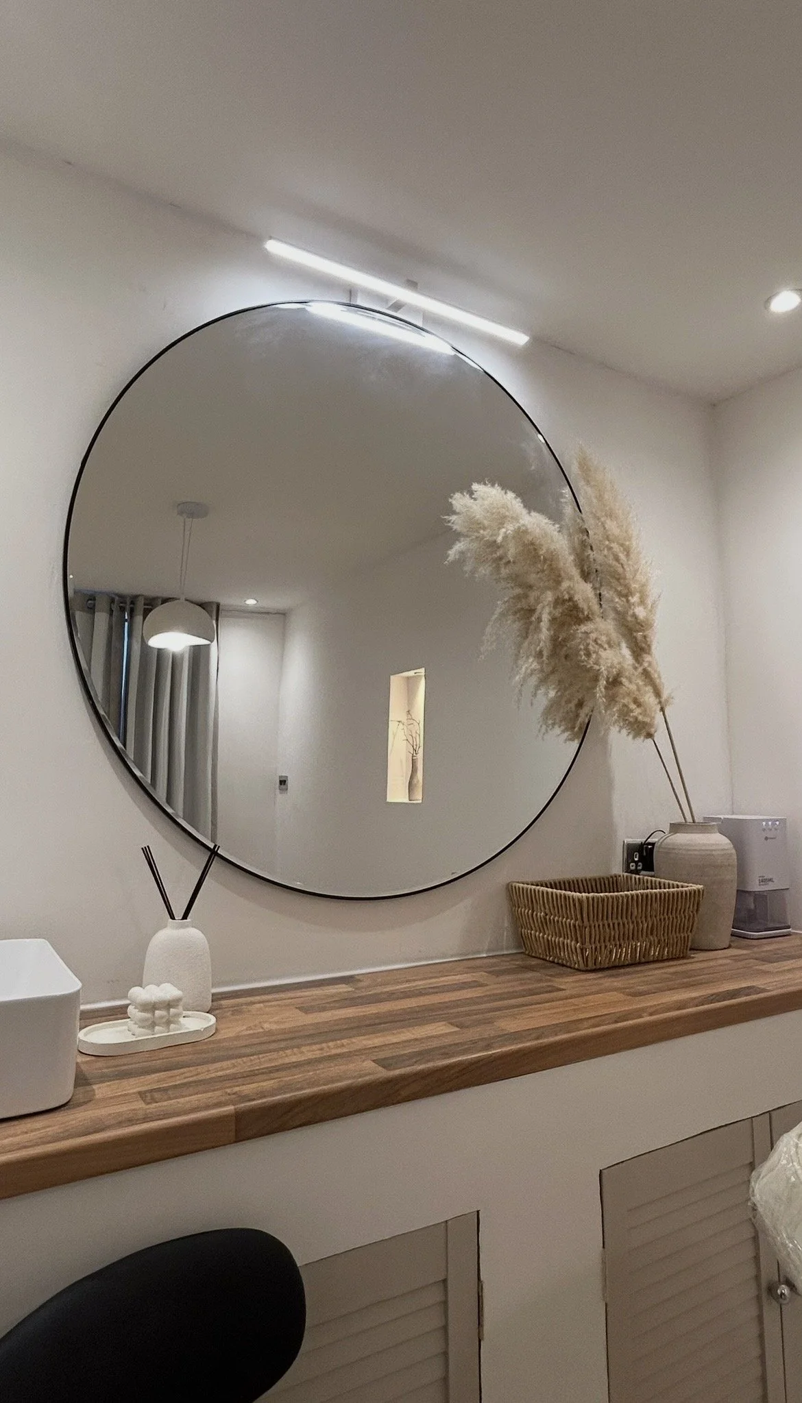A mirror hung on a wall reflecting ceiling lights, a hanging lamp, and window with curtains. On the wooden surface below are decorative items including pampas grass in a container, a small white vase with reed sticks, and a woven basket.