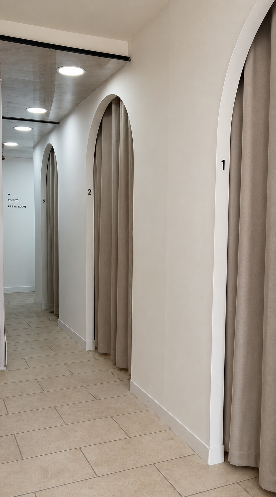 A corridor with three fitting rooms numbered 1, 2, and 3 with beige curtains, white walls, and beige tiled flooring. In the background, there is a sign pointing to a toilet and break room.