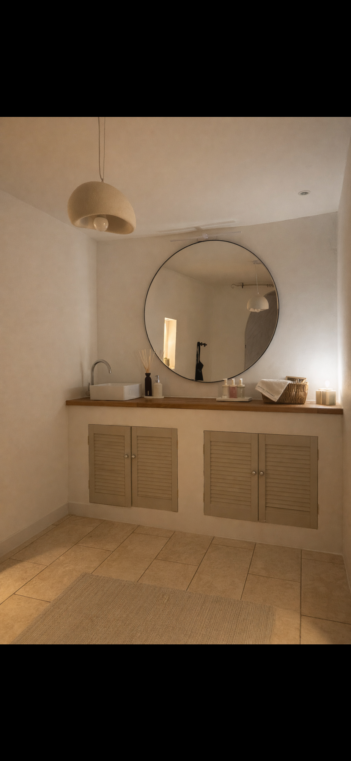 A bathroom vanity with a round mirror, a sink, and various toiletries and decorative items on the wooden countertop, with a beige tiled floor and neutral-colored cabinets below.