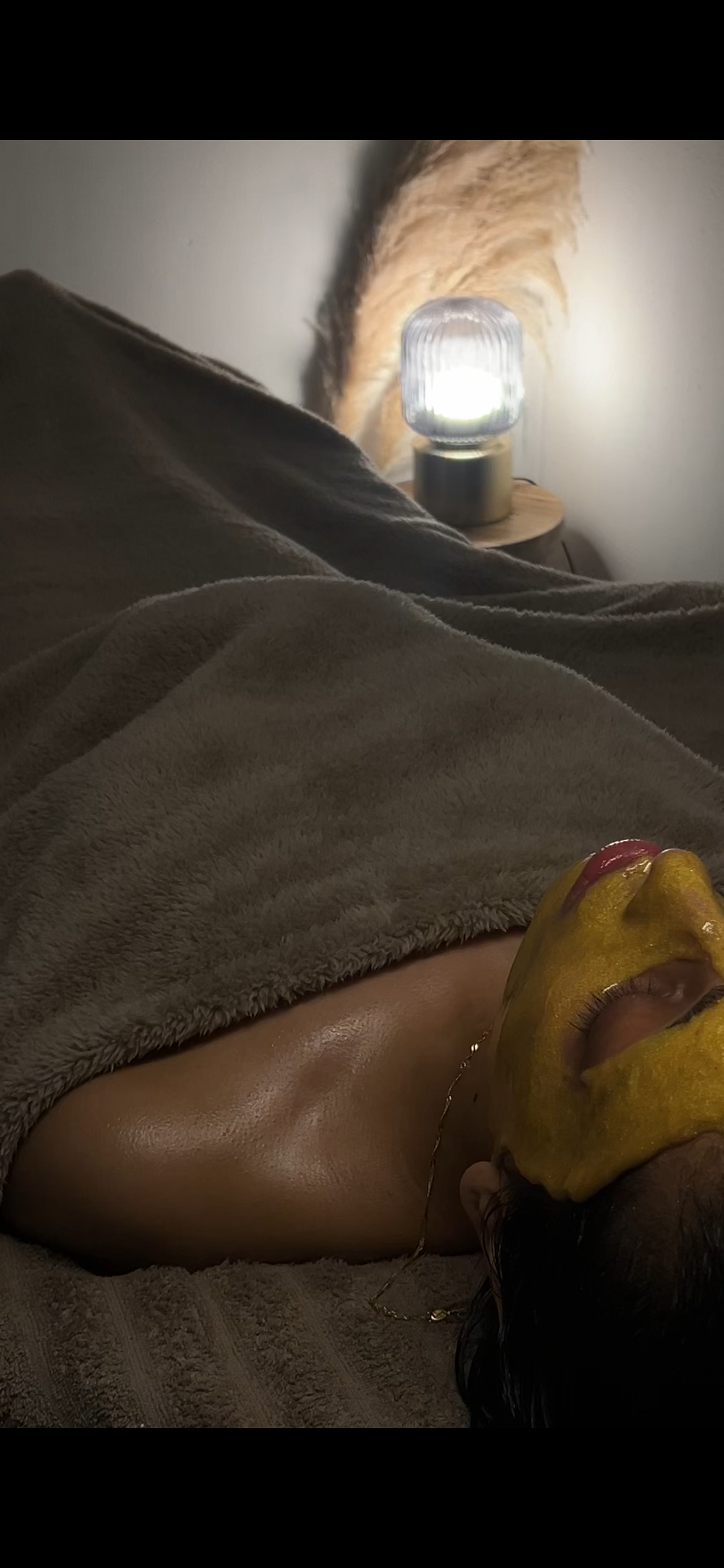 Person lying on bed covered with a grey blanket, wearing a yellow facial mask, with a gold chain around the neck. In the background, there is a wooden nightstand with a lit lamp and a feather decoration.