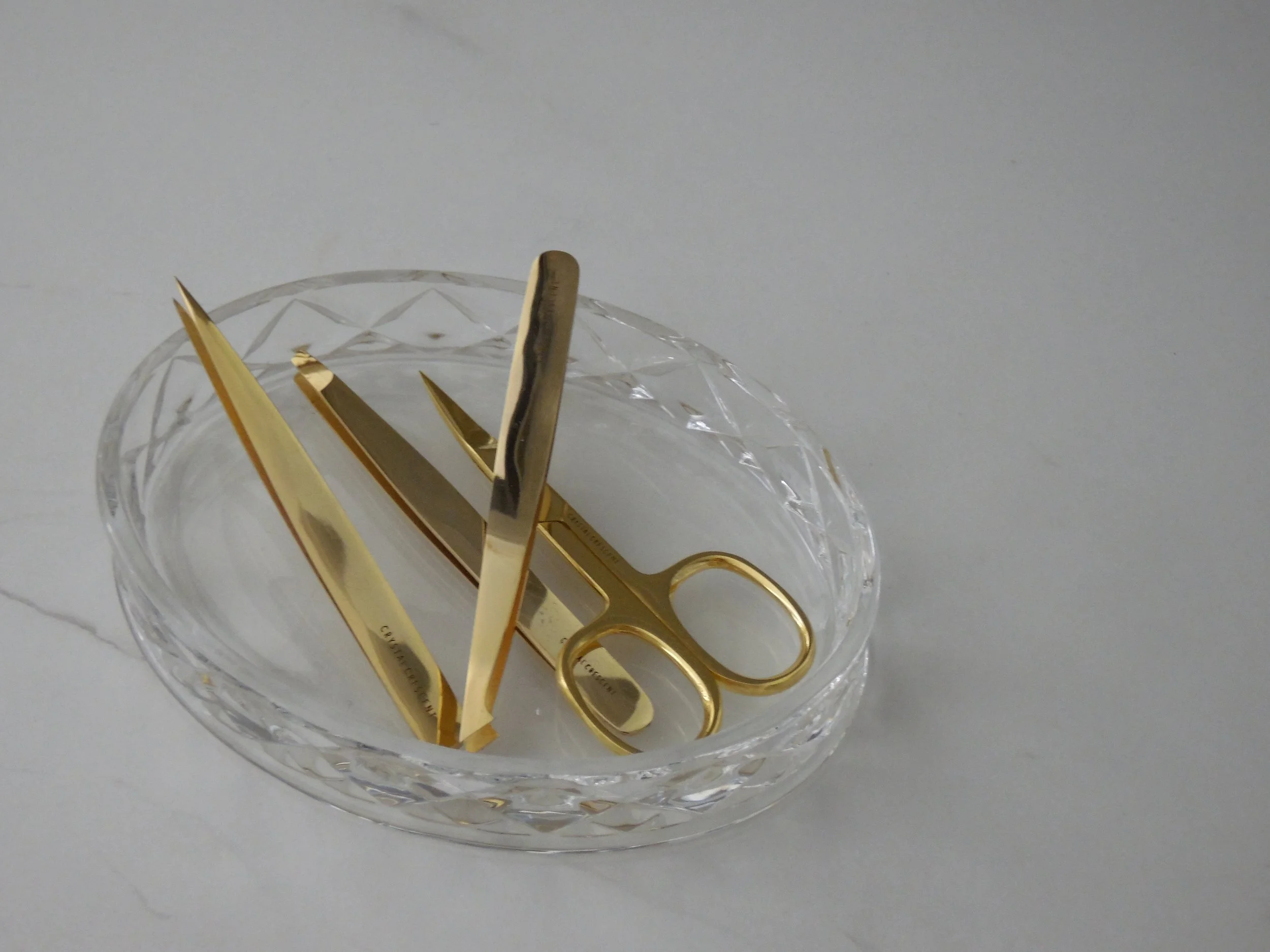 A glass dish holding gold-colored scissors and nail clippers on a white surface.
