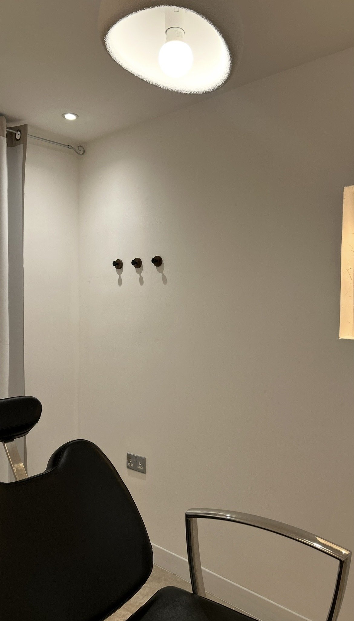 Interior view of a room with a ceiling light, three hooks on the wall, an electrical outlet, a black chair, and part of a curtain on the left side.