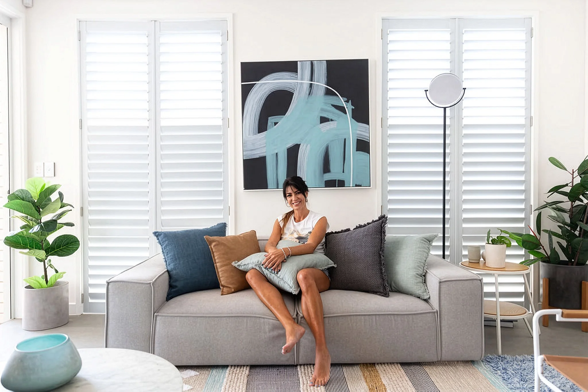 A woman sitting on a light grey couch smiling, with her legs crossed and her arms resting on their lap. She is surrounded by various throw pillows, with windows and white plantation shutters in the background. There are green plants on both sides of 