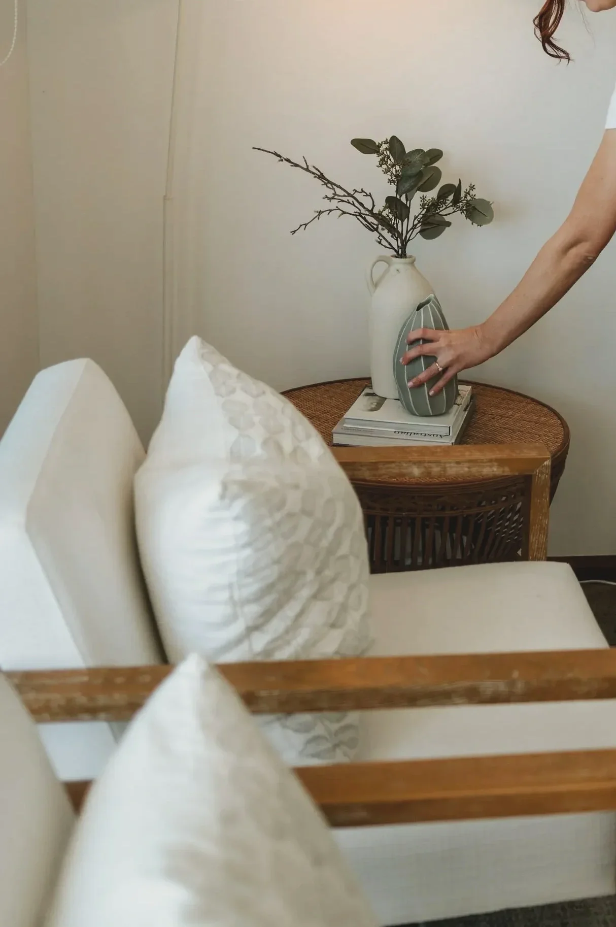A table with a vase of greenery, a stack of books, and a person placing a decorative object on the table, in a cozy, well-lit room.