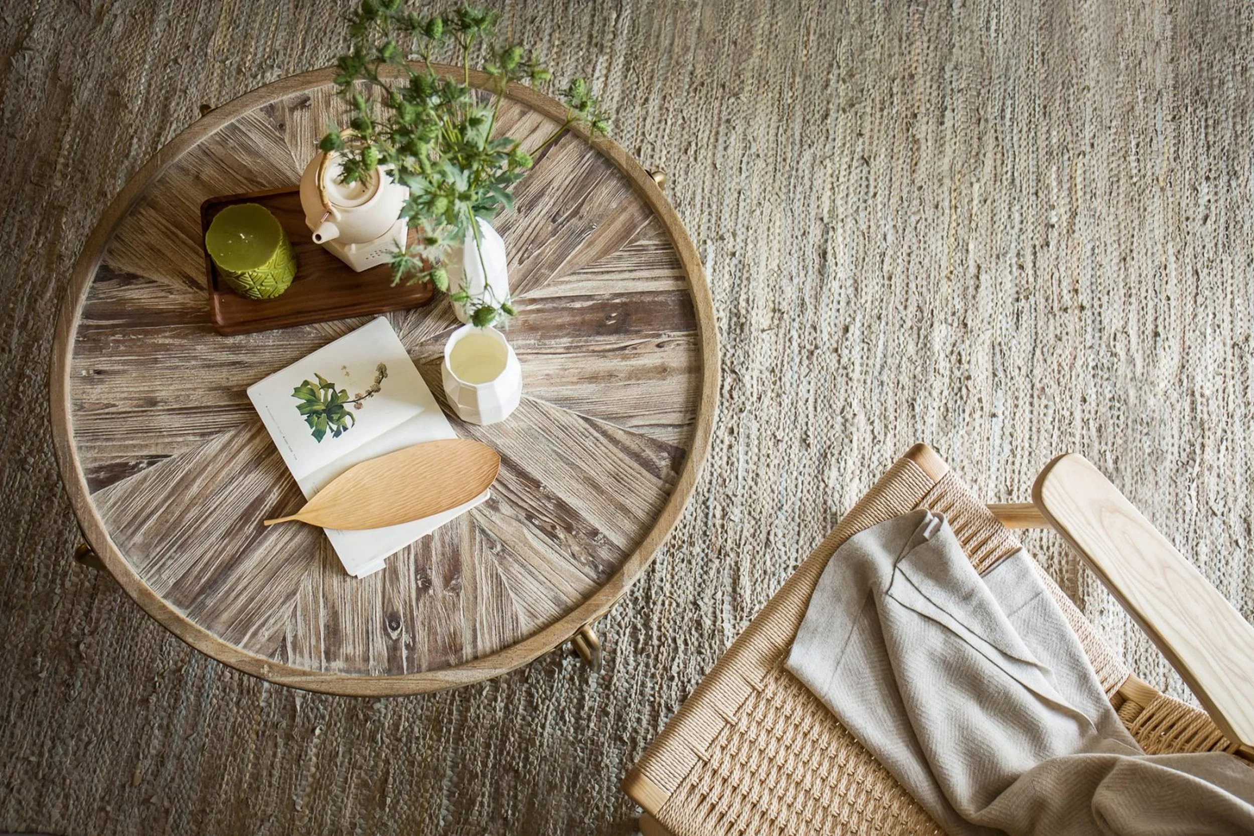 Round wooden coffee table with a white teapot, a small plant in a white vase, a green candle, a white cup, and a plant illustration on paper, set on a textured beige carpet.
