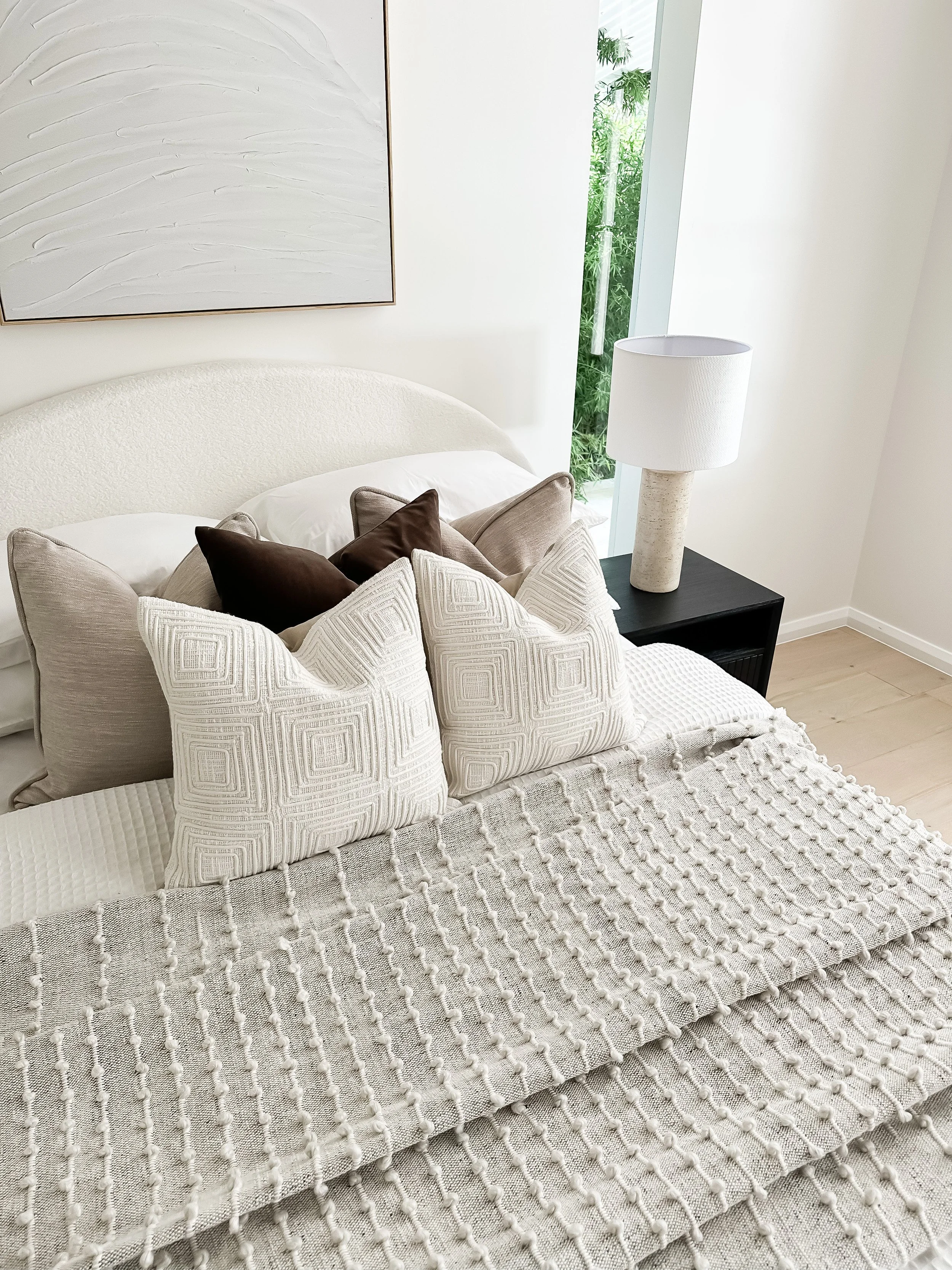 A living room corner with a white textured armchair with patterned pillows, a black side table with a white lamp, a large abstract painting, and a window revealing greenery outside.