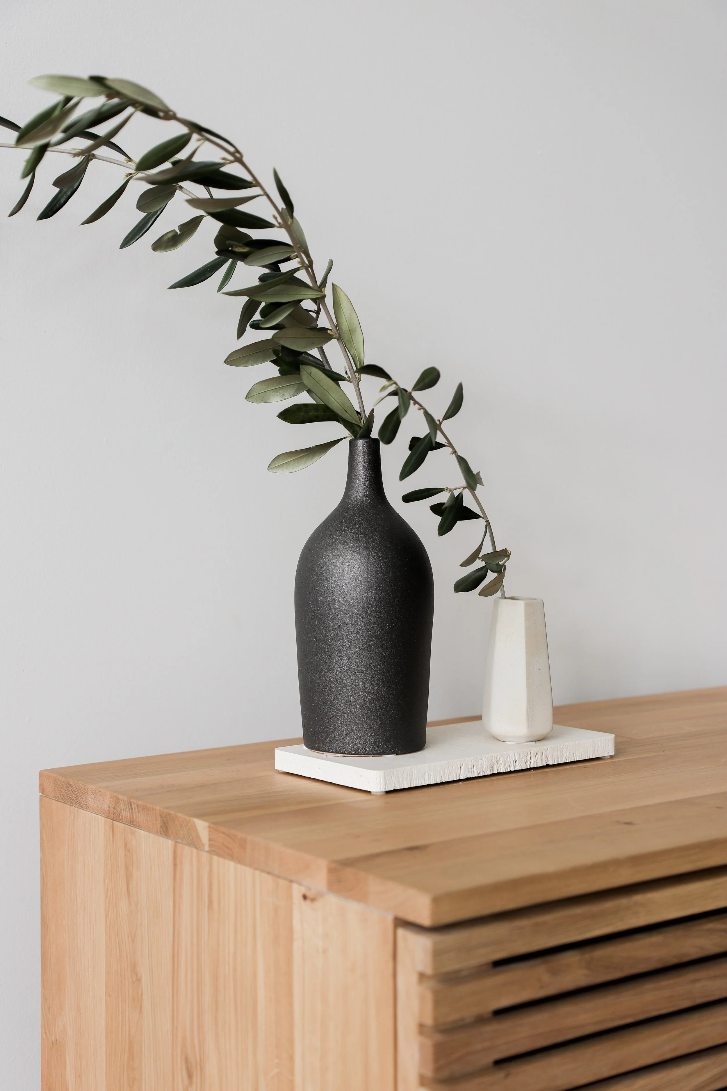 Two vases with greenery, one black and larger, the other white and smaller, on a wooden surface with a white textured platform underneath.