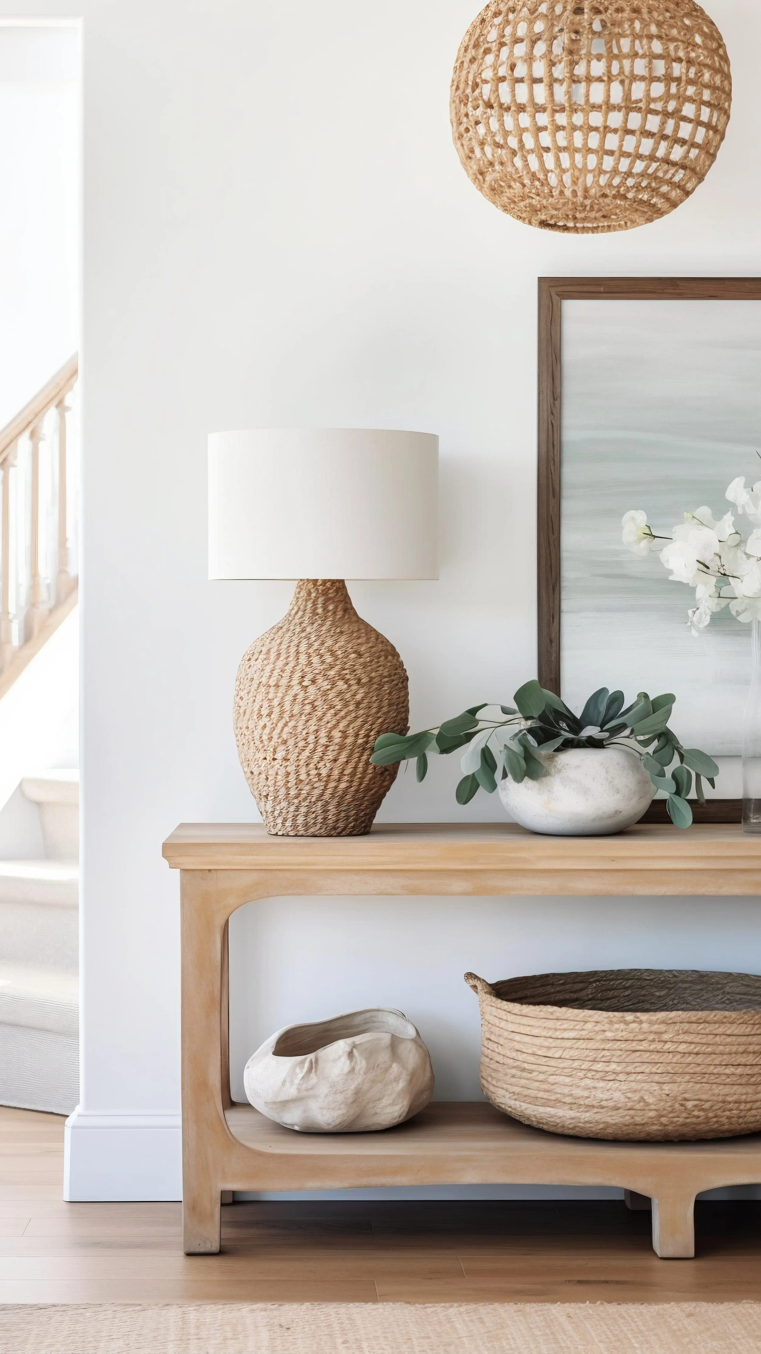 A white wall with a wicker hanging light, a table lamp with a woven wicker base, a picture frame with light-colored streaks, white orchids in a white pot, and decorative woven baskets and a ceramic sculpture on a light wooden console table.