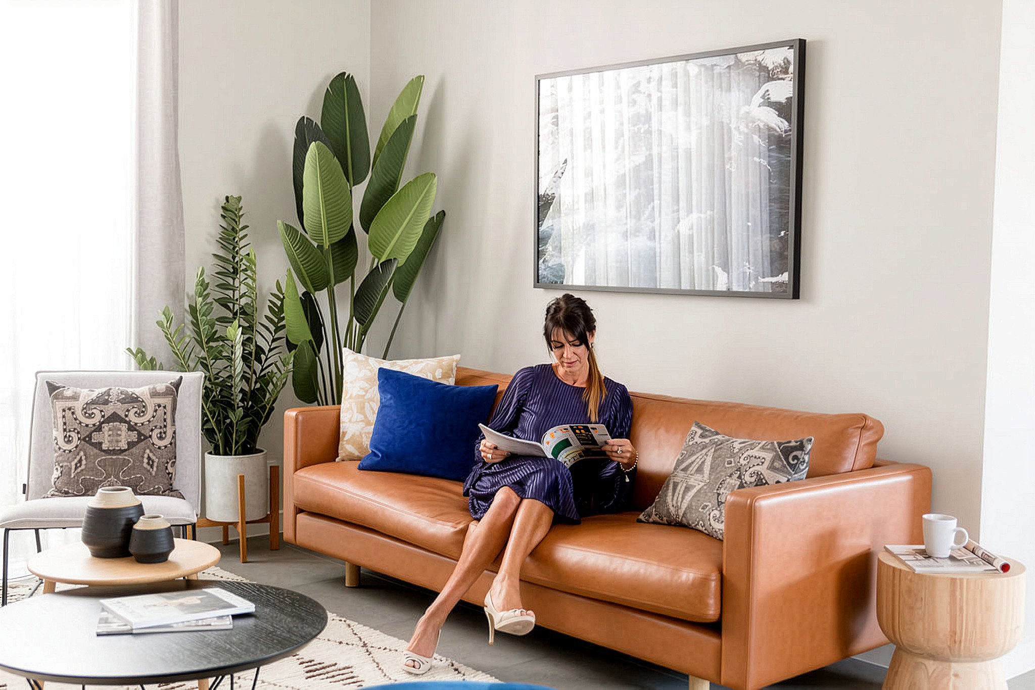 A woman sitting on a tan leather sofa with a blue pillow, reading a magazine in a modern living room with plants, a wall art picture, and a small wooden side table.