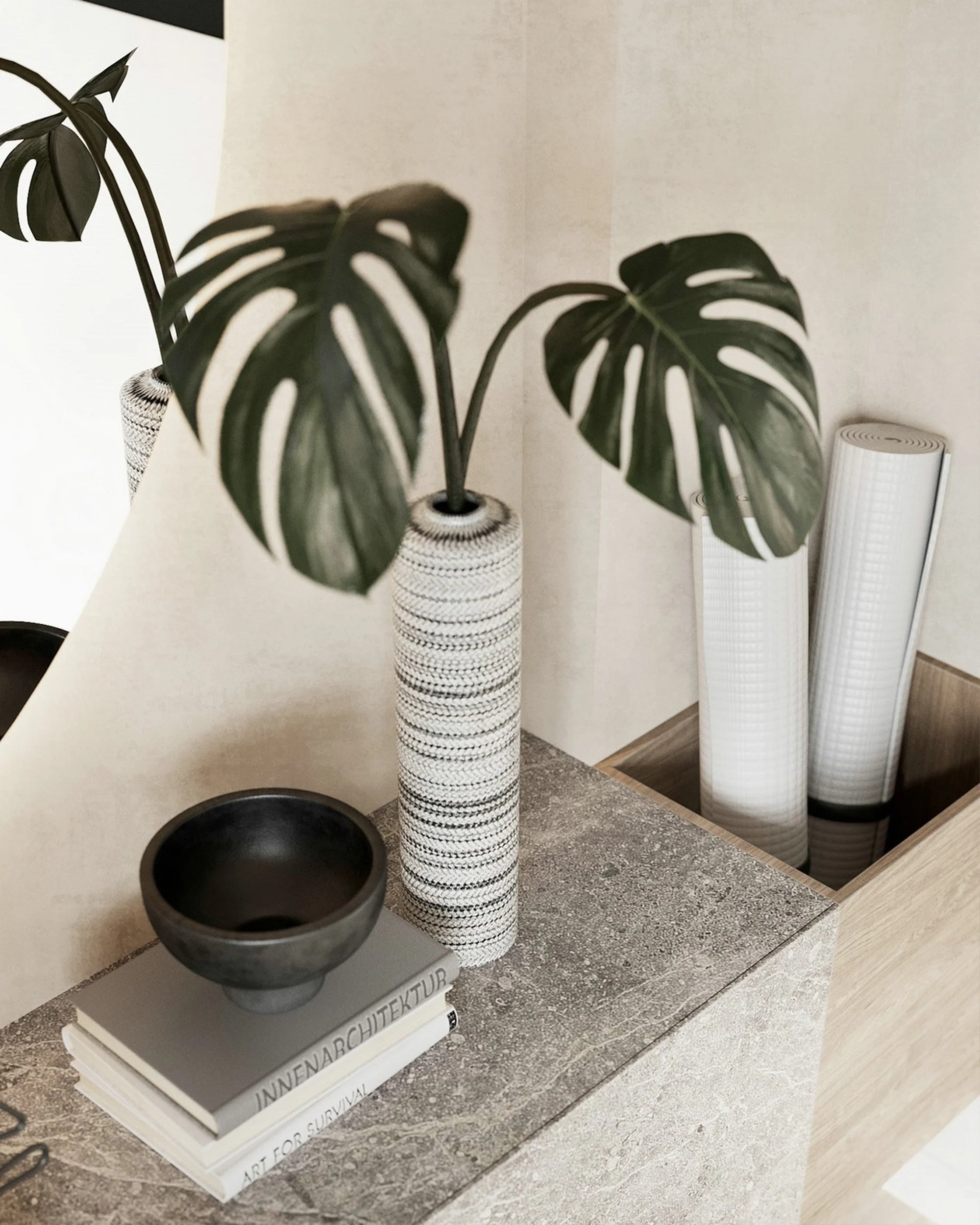 A black ceramic bowl on a white display stand, with three rolled-up white textured mats and a tall vase with a tall houseplant with split green leaves in the background.