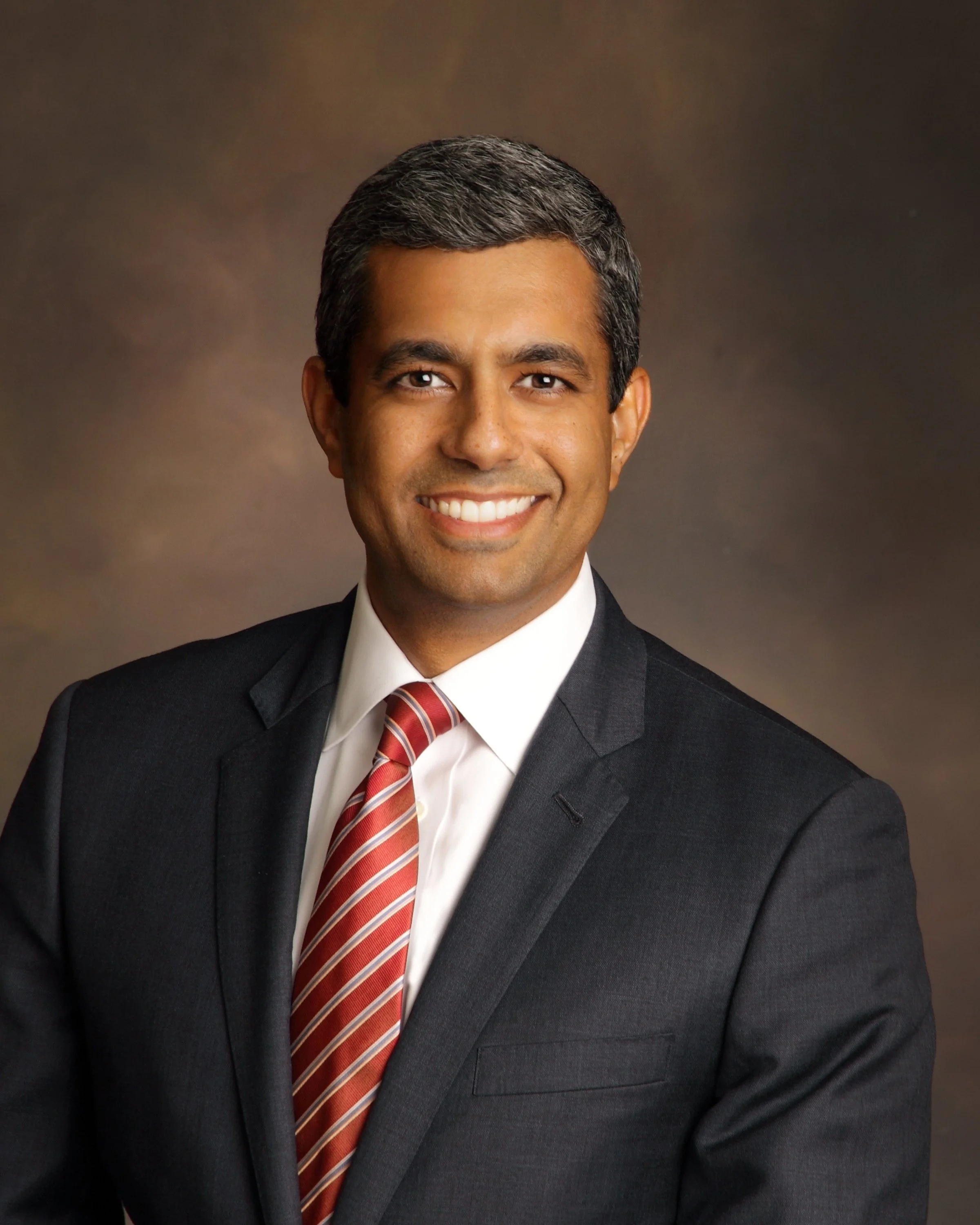 Professional headshot of David Chevalier in a black suit and striped red tie, smiling against a brownish background.