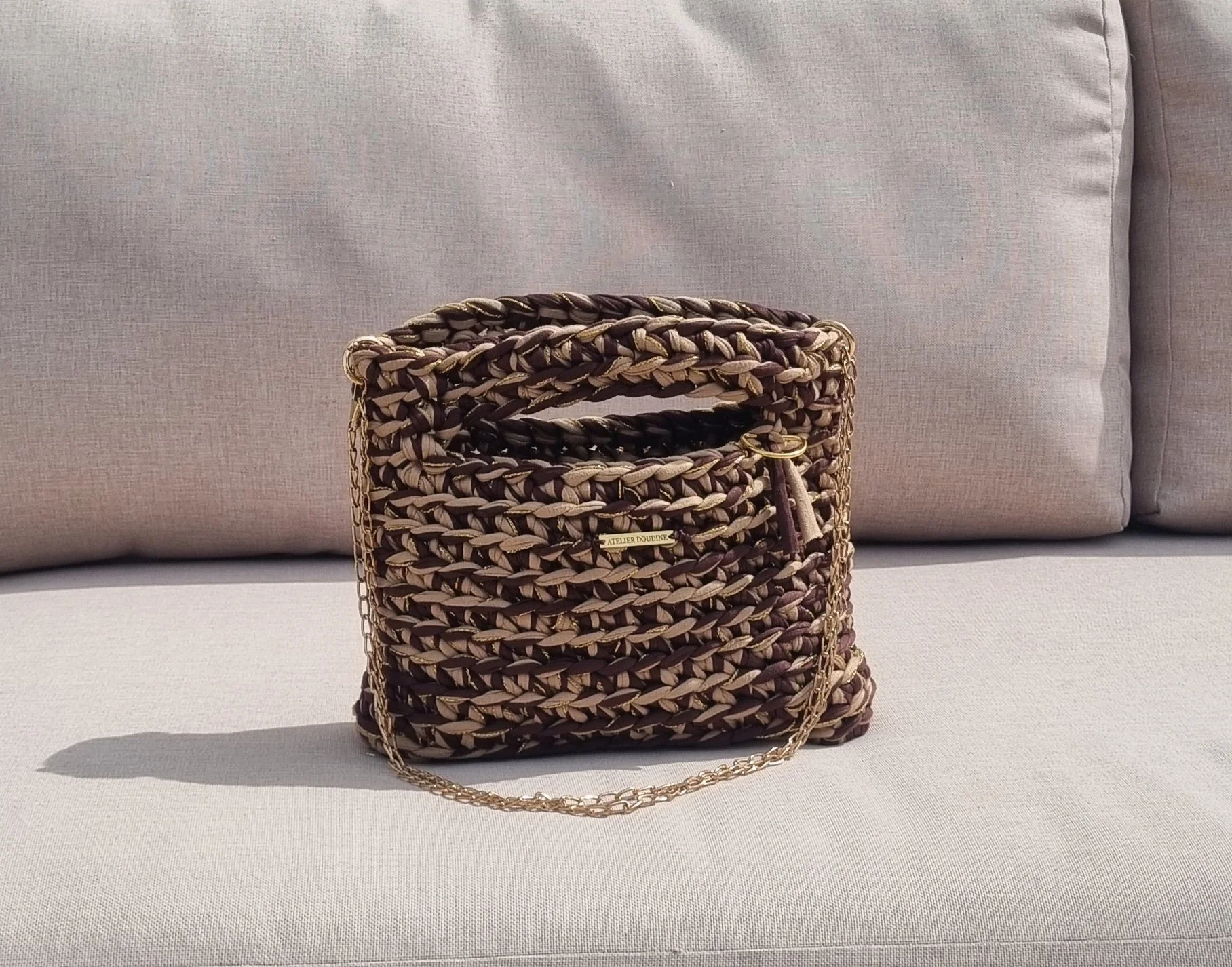 Sac à main crocheté de couleur marron avec chaîne en or, posé devant un canapé en tissu beige.