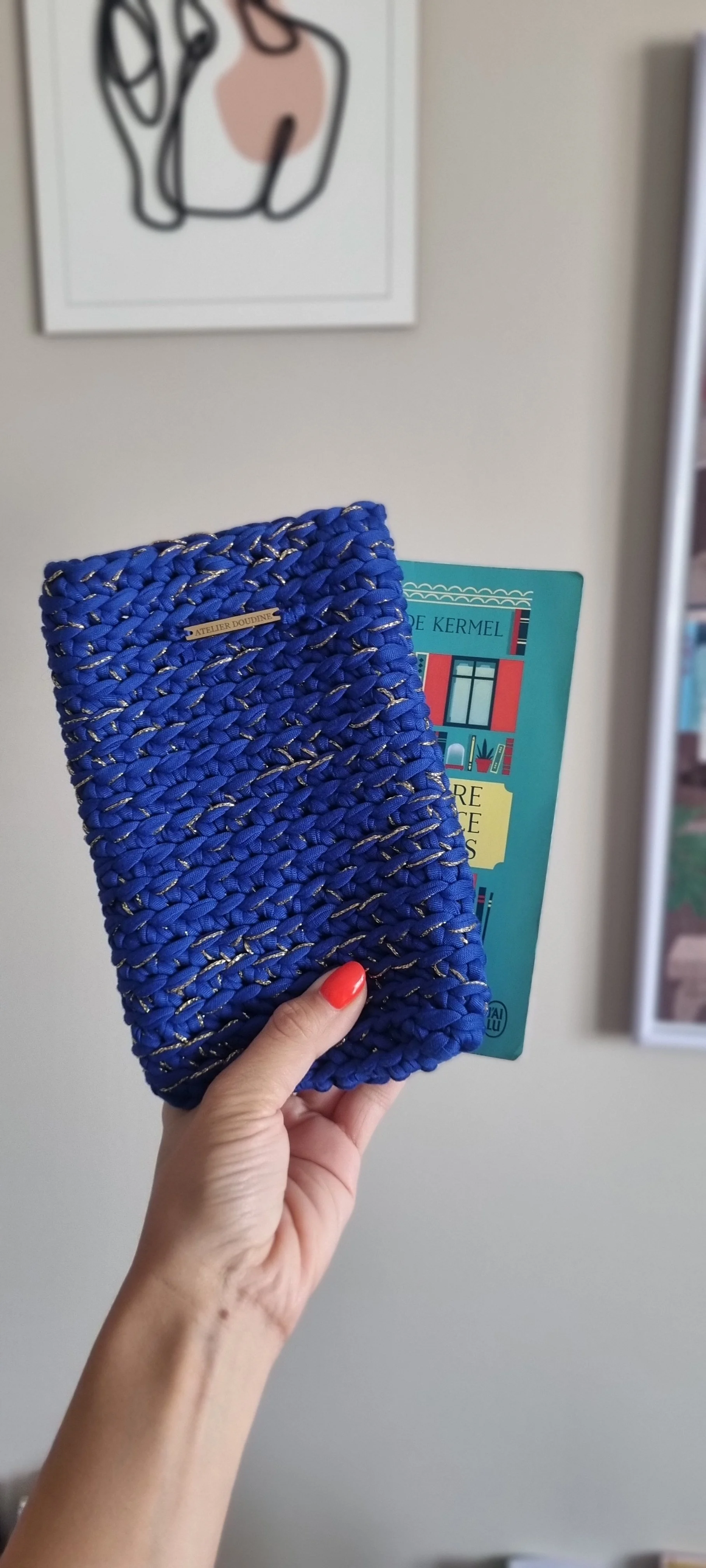 Main tenant une pochette à livre bleue en tissu tressé, avec une étiquette portant la mention "ATELIER DOUDINE".
