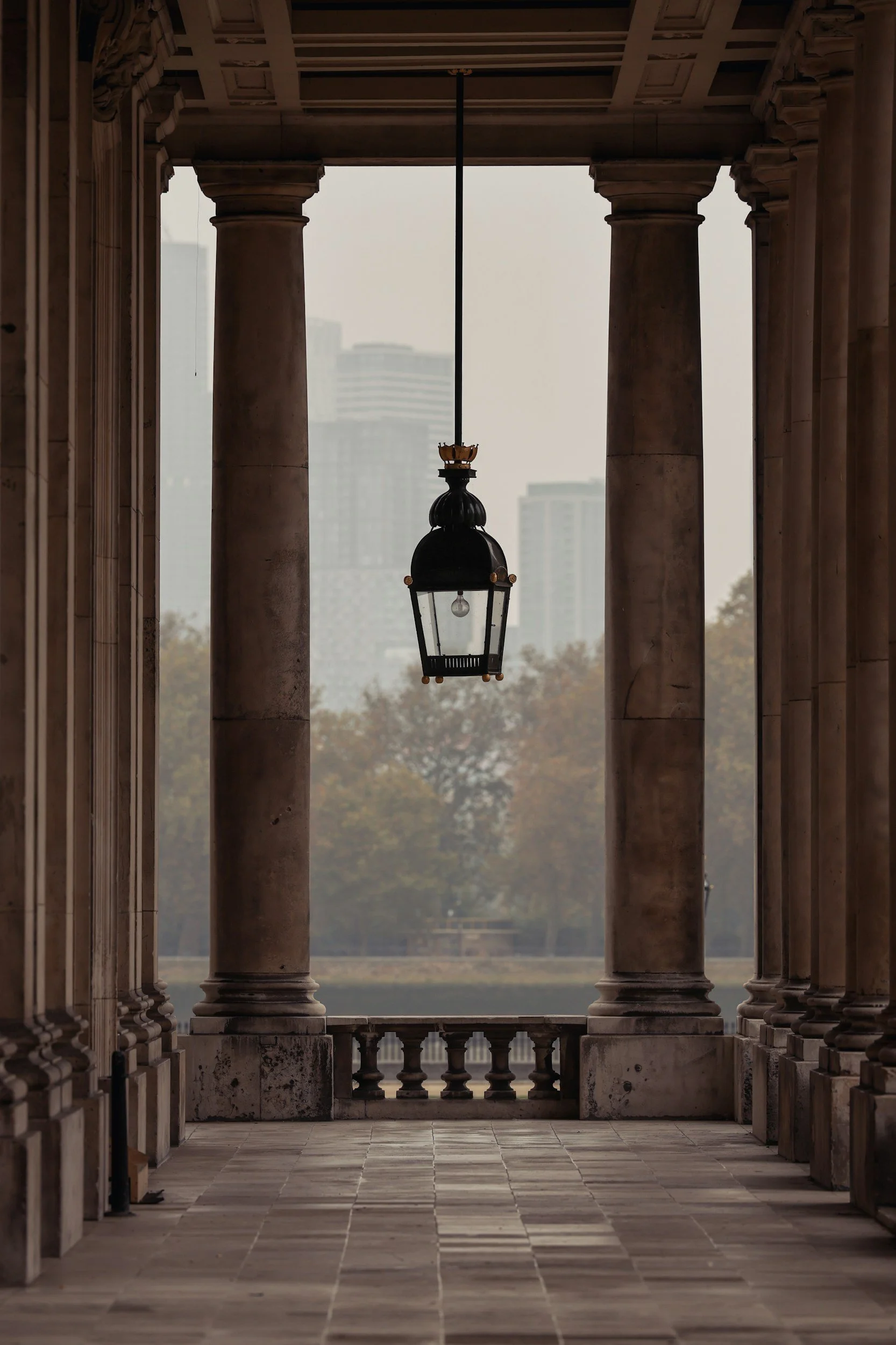 A covered walkway with stone columns and a balcony railing, featuring a hanging lantern in the center, overlooking a misty cityscape with tall buildings in the background.