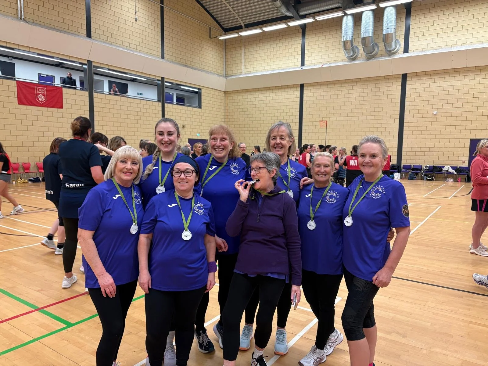 Group of women wearing blue sports jerseys and medals posing inside a gymnasium, celebrating a sports event.