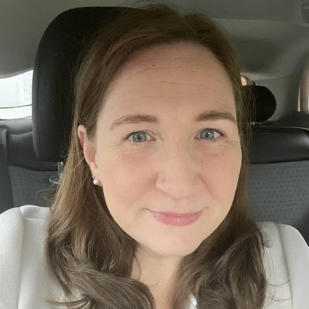 A woman with light skin, long brown hair, and blue eyes, smiling while sitting in a car.