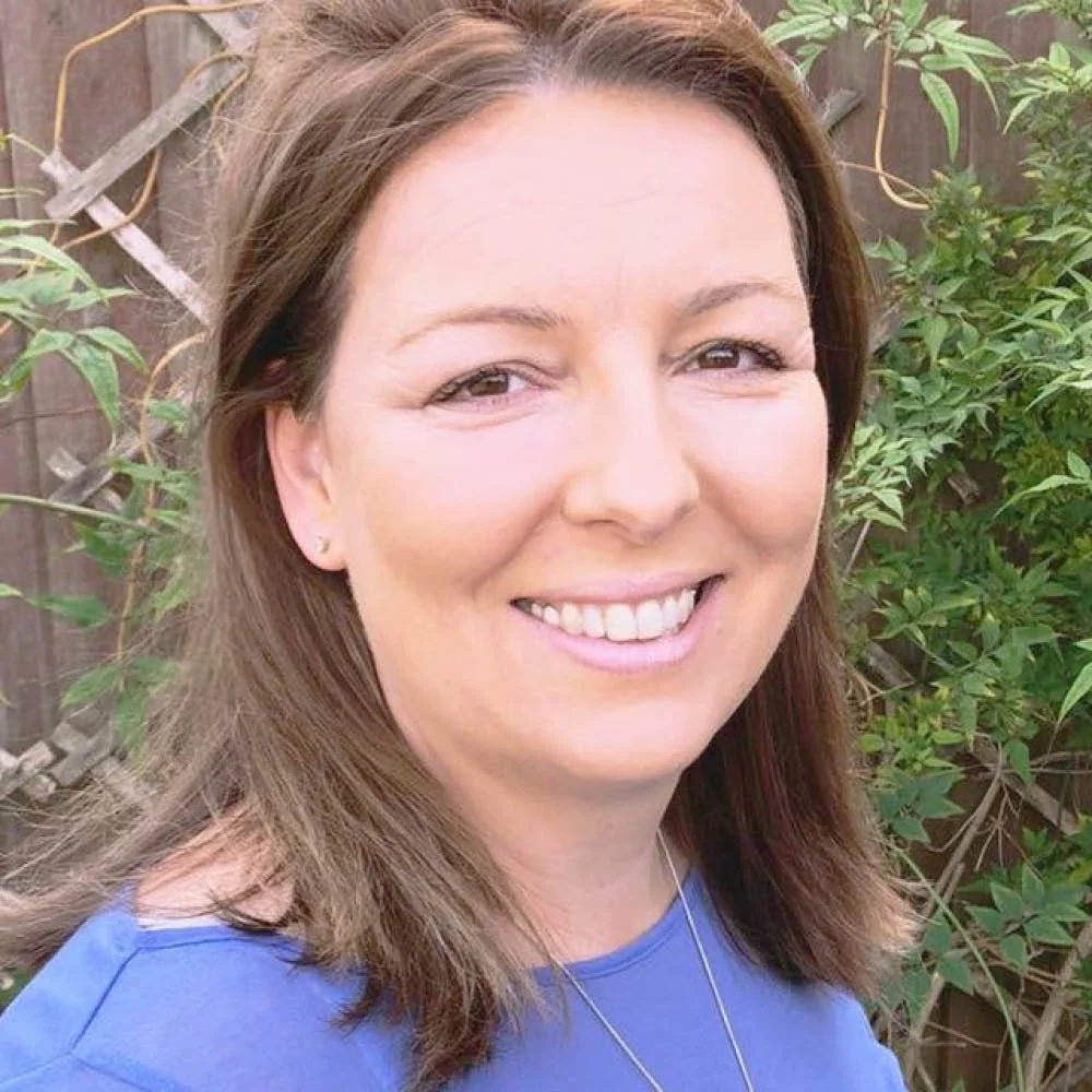 Close-up of a woman smiling outdoors, with a background of green foliage and a wooden garden fence.