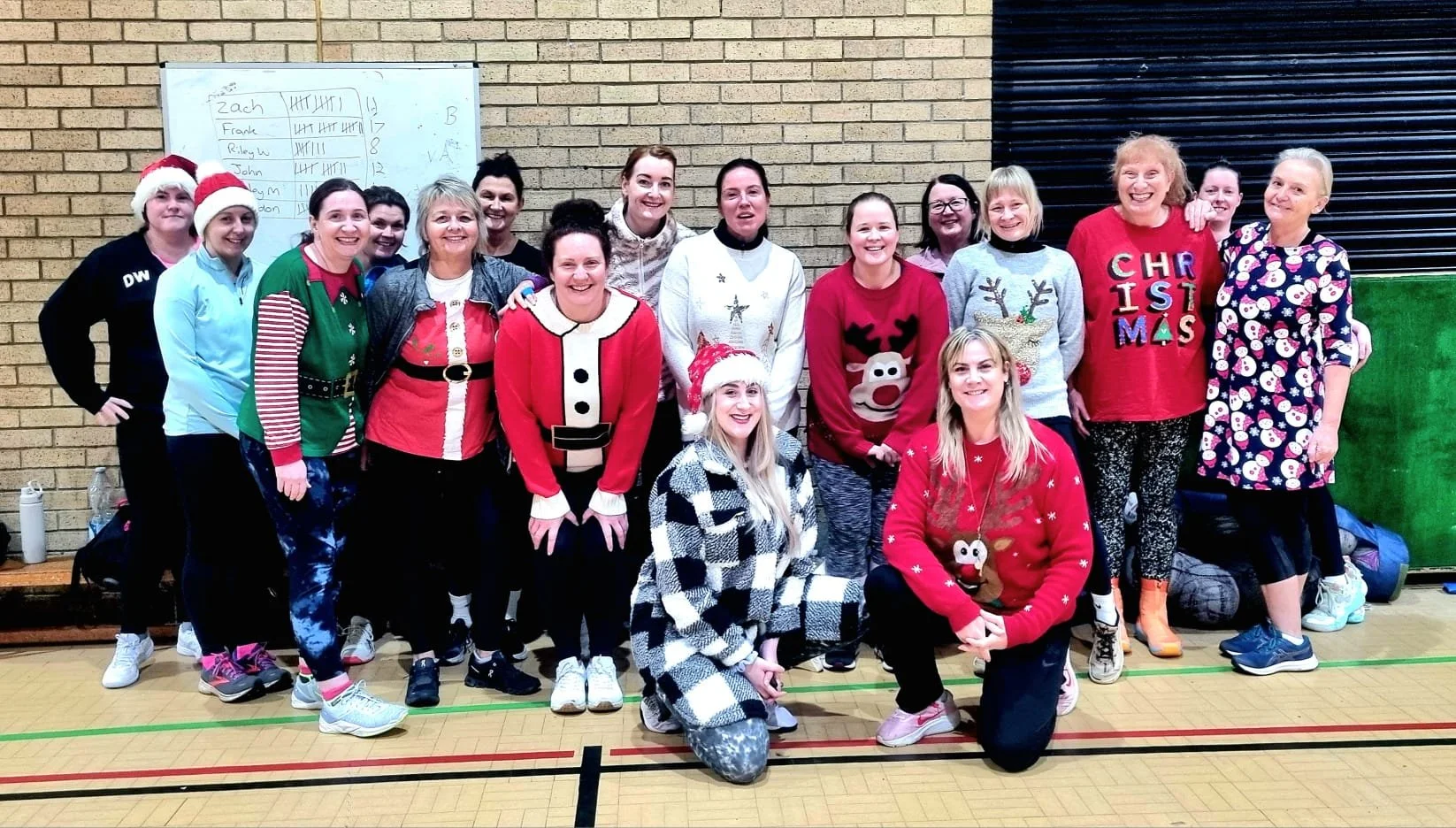 Group of women celebrating Christmas in a gym, wearing holiday-themed sweaters and hats, some with reindeer, Santa, and Christmas messages, standing in front of a brick wall and a whiteboard with tallies and names, smiling and posing for the photo.