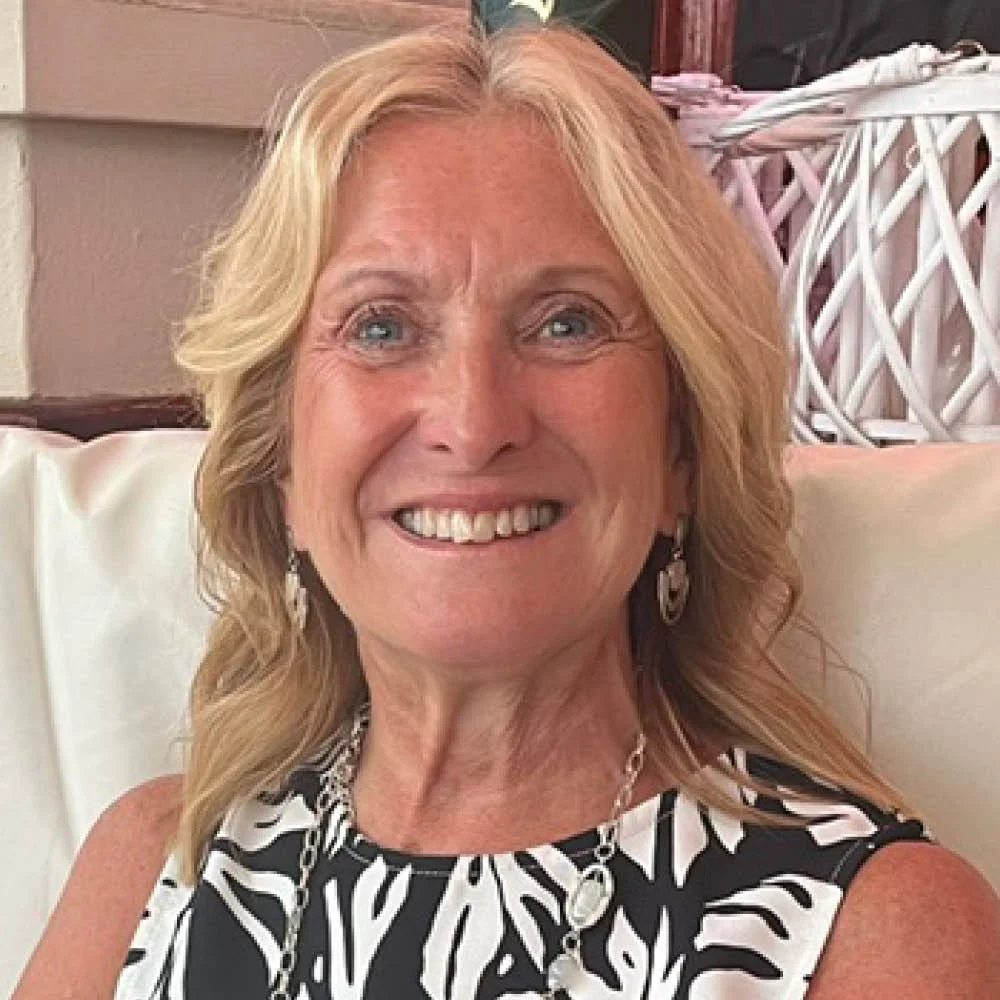 A smiling woman with blonde wavy hair wearing a black and white patterned sleeveless top, a necklace, and earrings, sitting on a white chair.