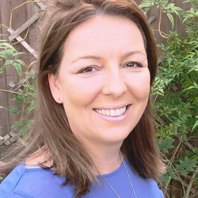 A smiling woman with shoulder-length brown hair wearing a blue top, standing outdoors in front of a wooden fence with green foliage.