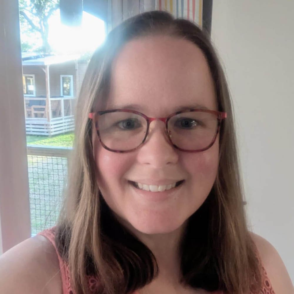 A woman with brown hair, glasses, and a pink top smiling indoors with a window and porch outside.