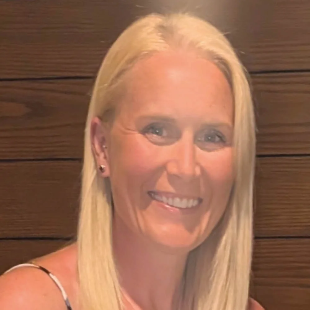 A woman with long blonde hair smiling, in front of a wooden wall background.