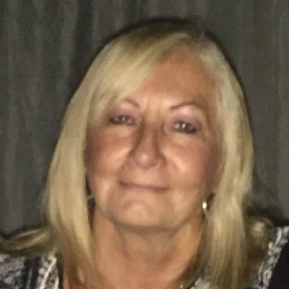 Close-up of a smiling middle-aged woman with blonde hair and earrings, against a dark background.
