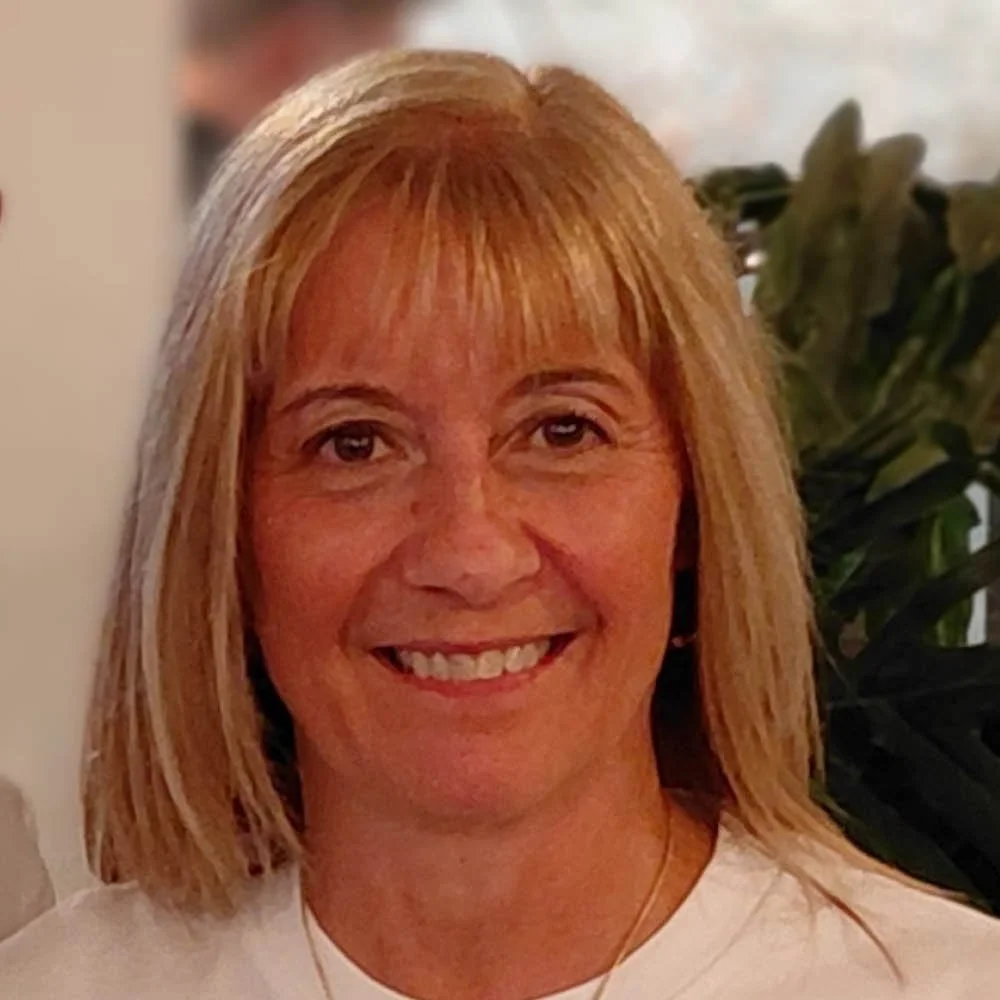 Smiling woman with blonde hair indoors, with green plants in the background.