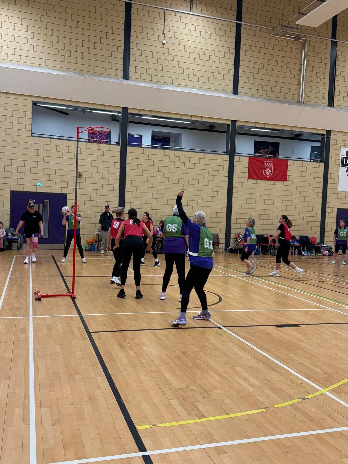 Women playing indoor volleyball in a gym, with a volleyball net and several players in red and black jerseys. Some players are wearing green and purple vests with 'GS' on the back. There are people on the side sitting on chairs and watching the game.
