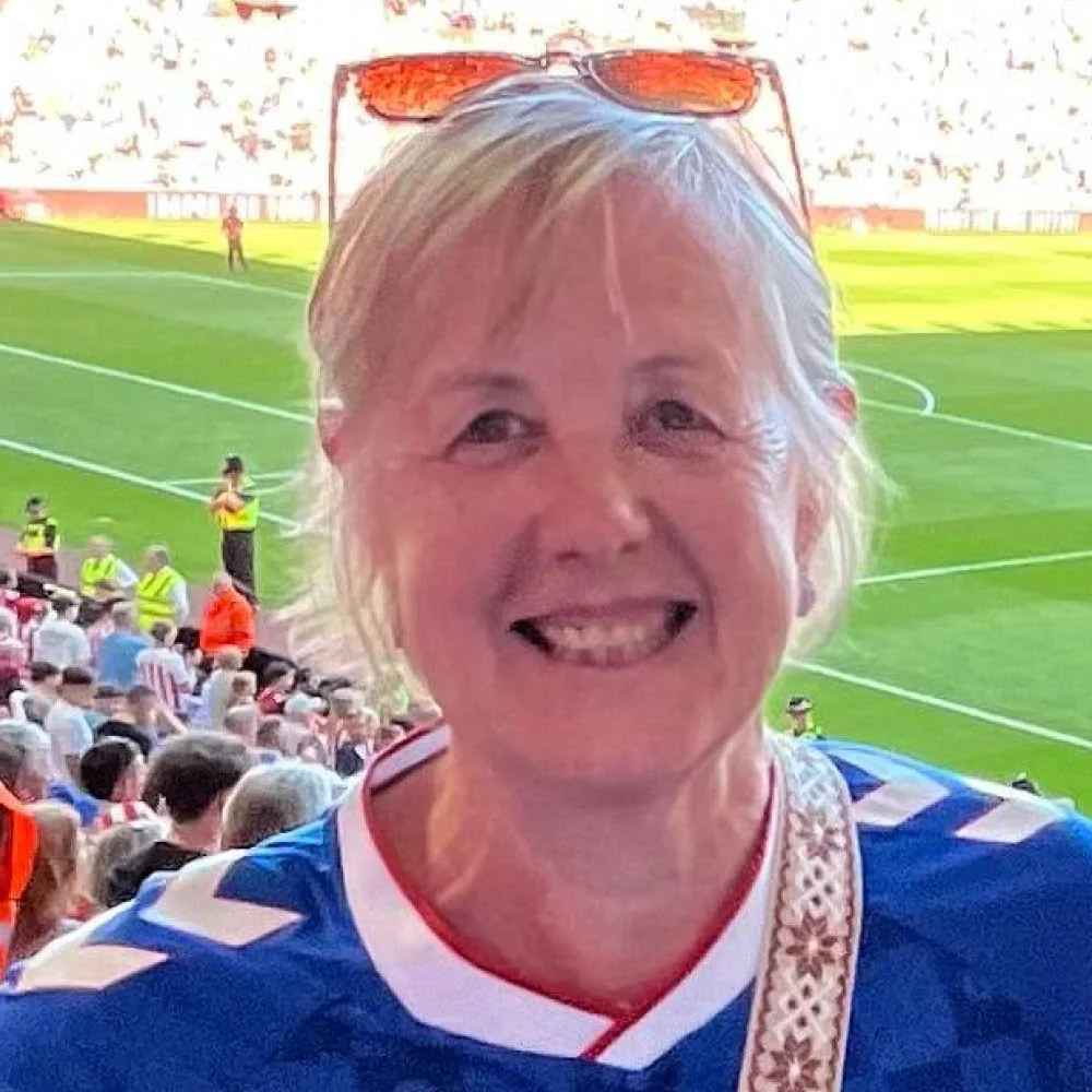 A smiling woman with blonde hair and sunglasses on her head at a soccer stadium, with a crowd and players on the field in the background.
