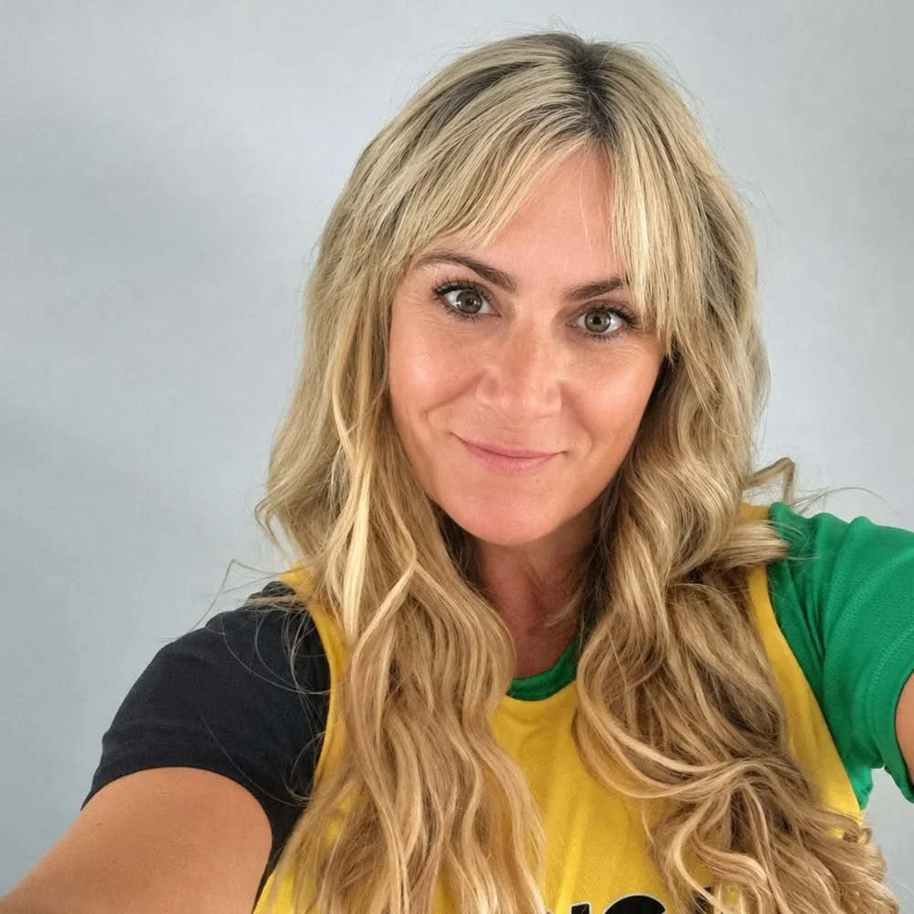 A woman with long wavy blonde hair and brown eyes, smiling, wearing a colorful shirt with green, yellow, and black sections, taking a selfie against a plain gray background.