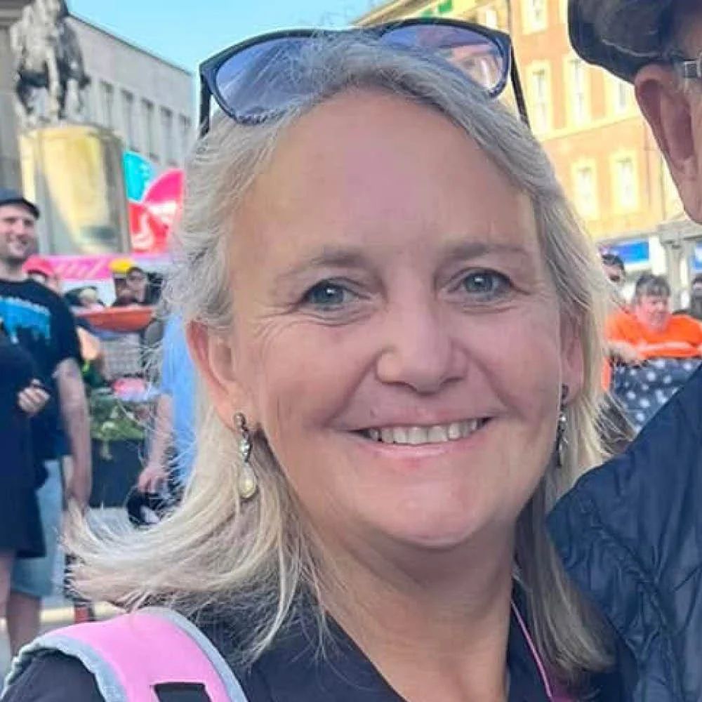 A smiling woman with blonde hair and glasses on top of her head. She is wearing earrings and a backpack, and is outdoors at a busy event or festival.