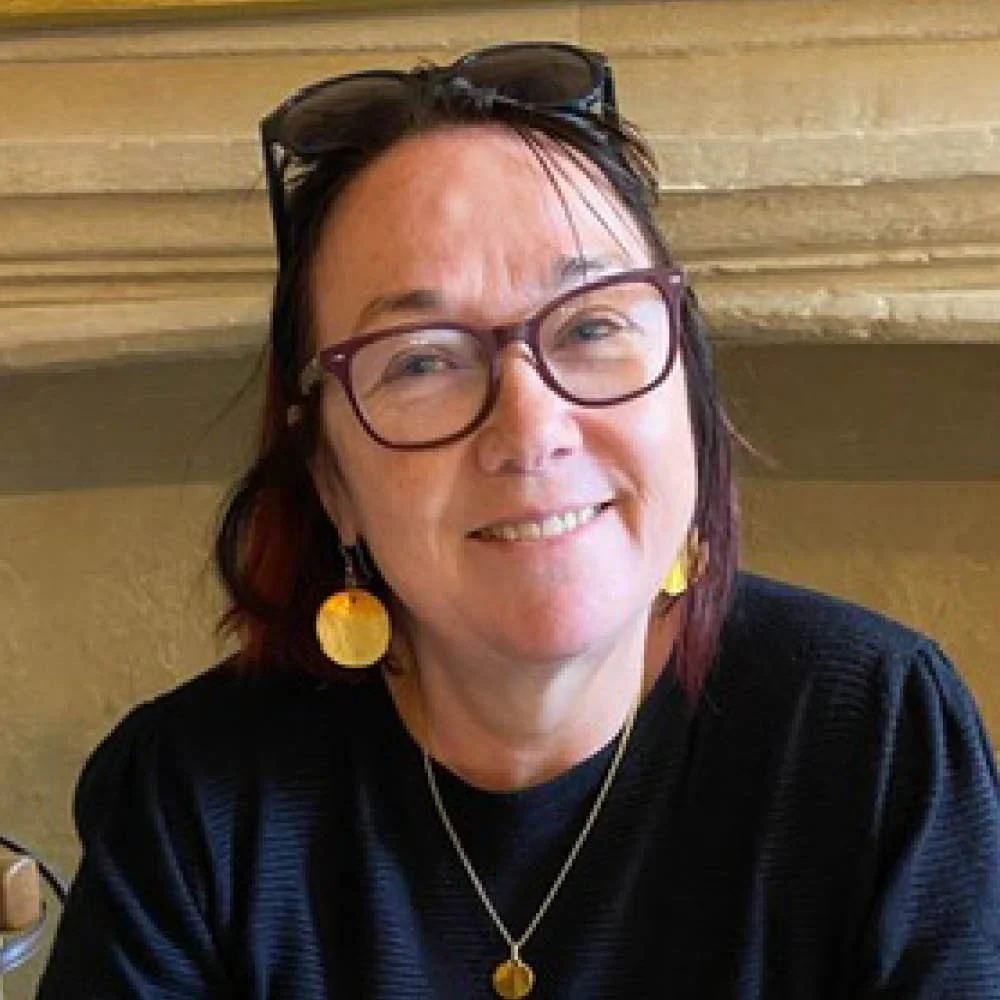 A smiling woman with glasses, shoulder-length dark hair, wearing earrings and a necklace, sitting indoors in front of a beige or tan brick wall.
