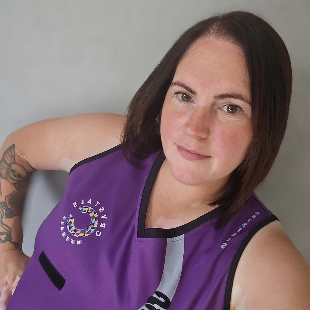 A woman with shoulder-length brown hair wearing a purple sports jersey with the Brighton Wildcats logo, standing against a gray wall.