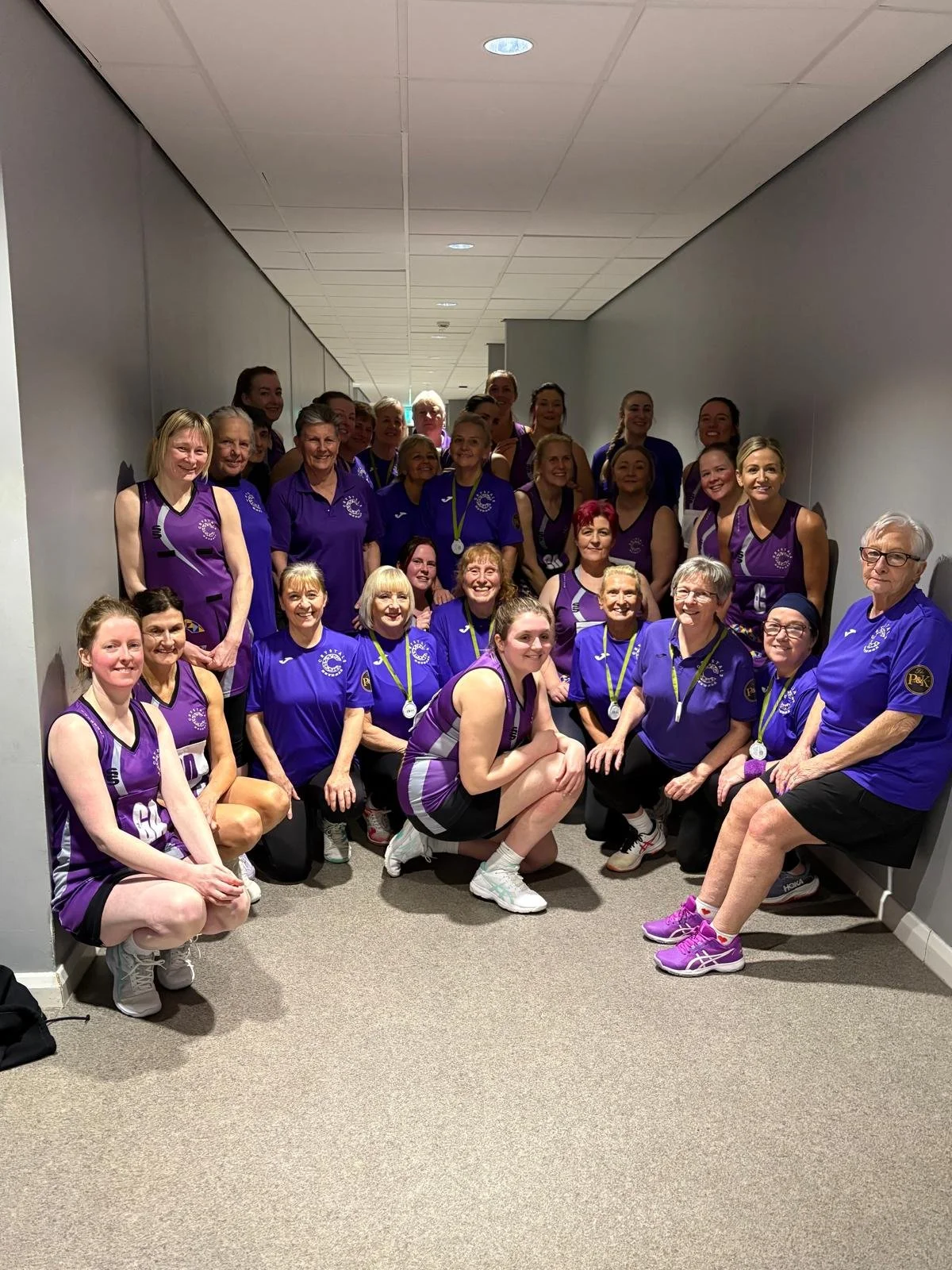 A large group of women in purple sports uniforms posing for a group photo in a hallway, some wearing medals around their necks, indicating they are part of a sports team or group.