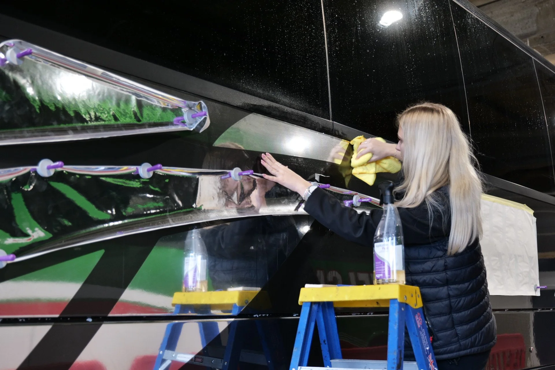 A woman polishing a black vehicle's exterior using a yellow cloth, with a blue and yellow ladder nearby and a clear spray bottle on top of the ladder.