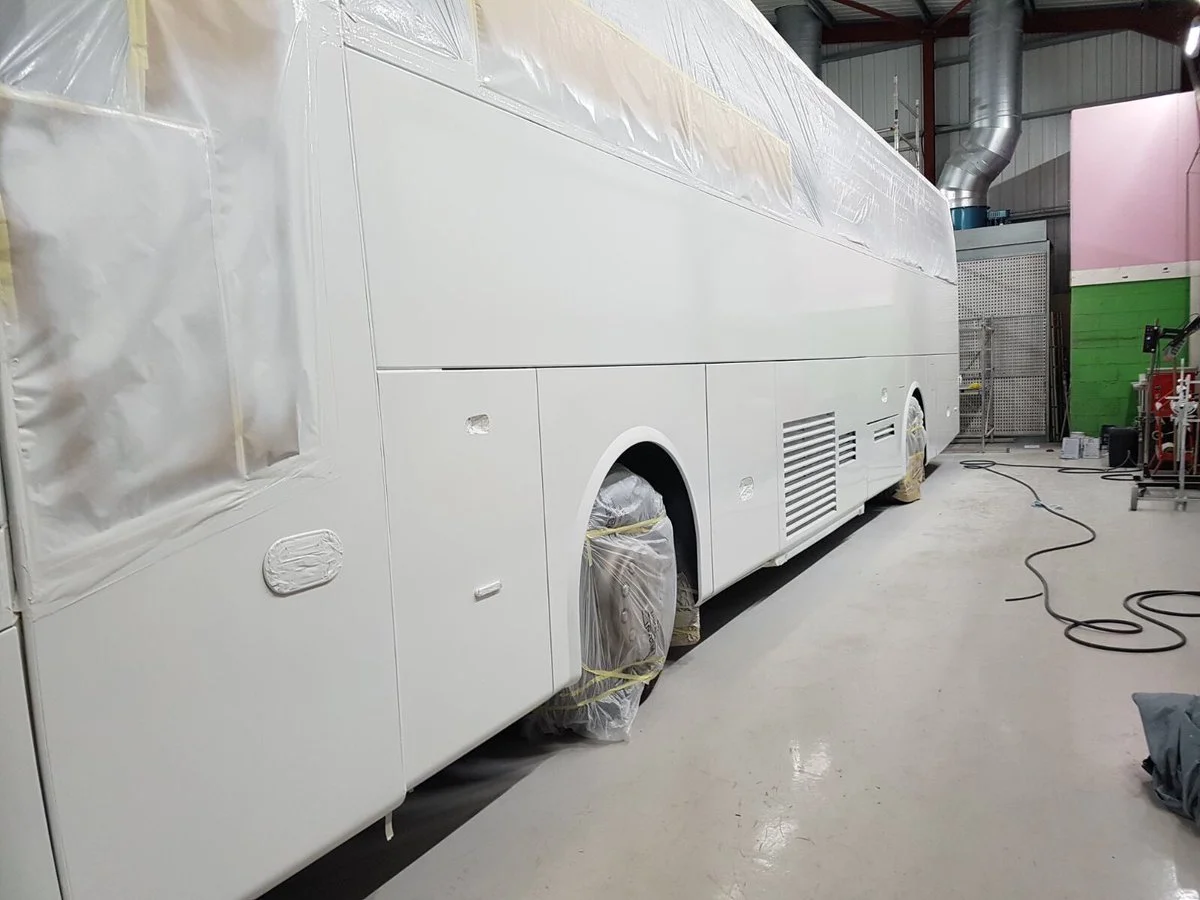 White bus inside a workshop, covered with masking paper and plastic, with wheels wrapped in protective plastic and tape.