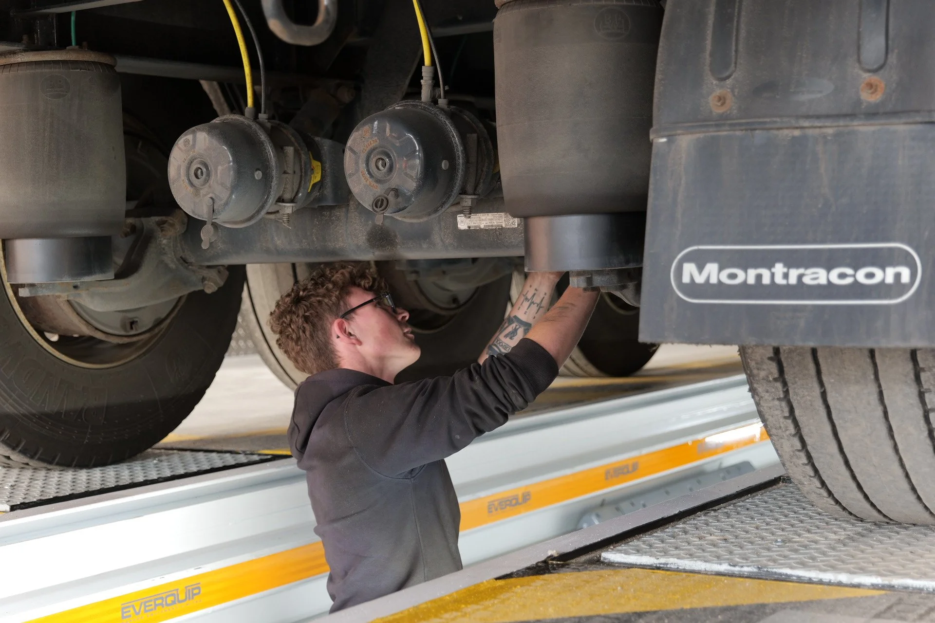 A mechanic working underneath a large truck, inspecting or repairing components, in a garage or workshop.
