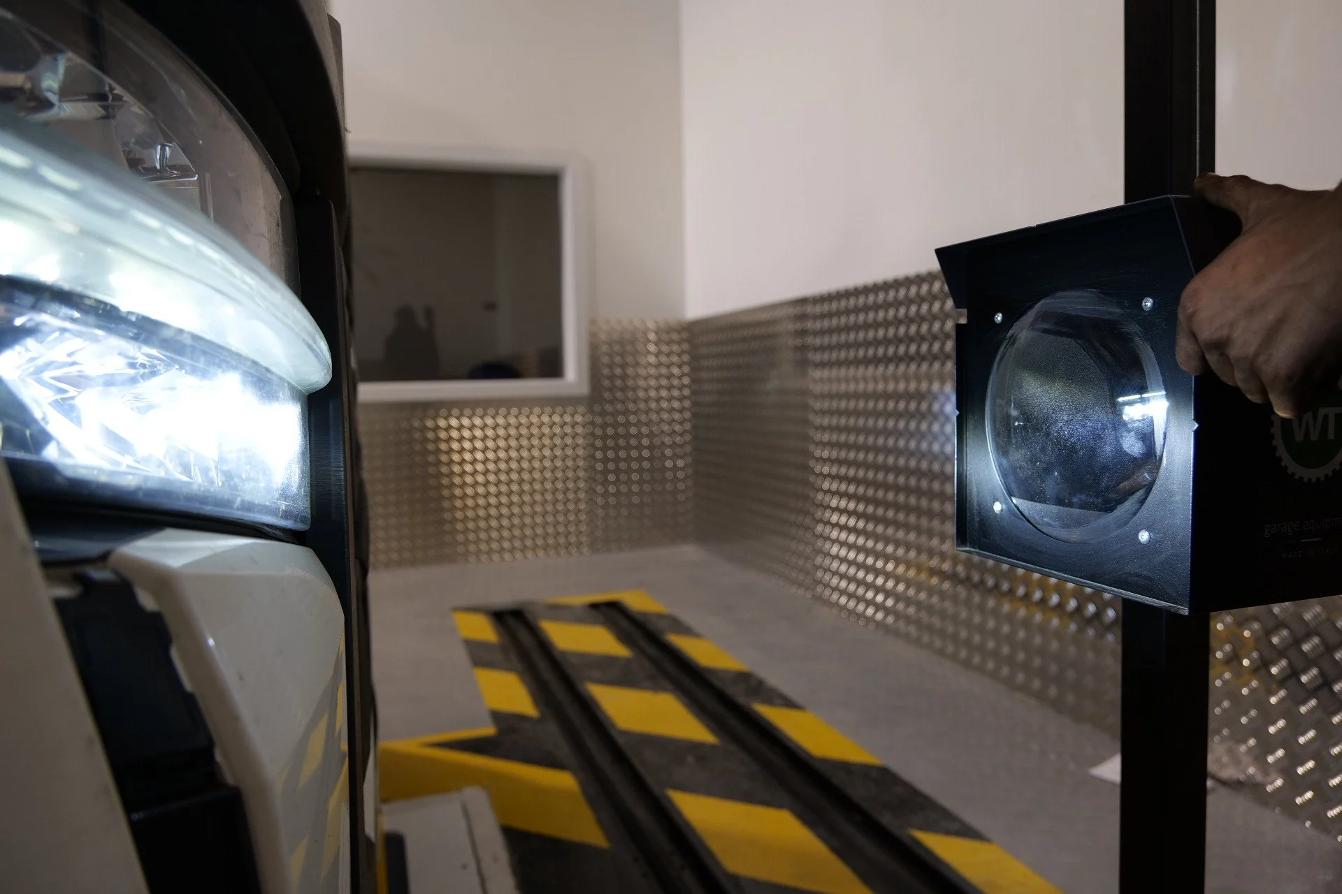 A person holding a black square device with a circular lens in front of a vehicle with a transparent cover, on a surface with yellow and black caution markings.