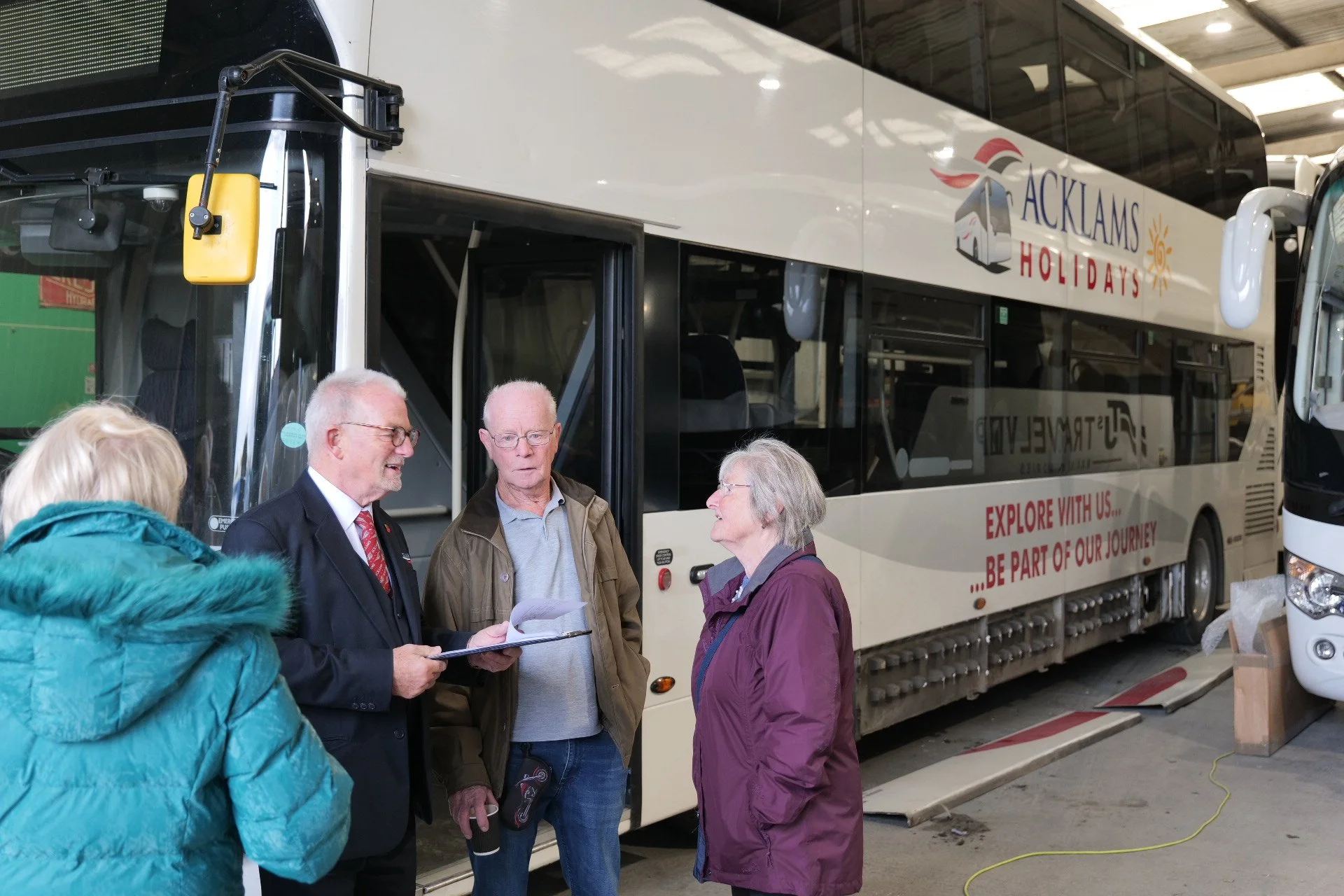 A group of four seniors standing in front of a double-decker bus inside a garage, engaging in conversation. The bus has 'Acklams Holidays' written on it and a slogan 'Explore with us... Be part of our journey'.