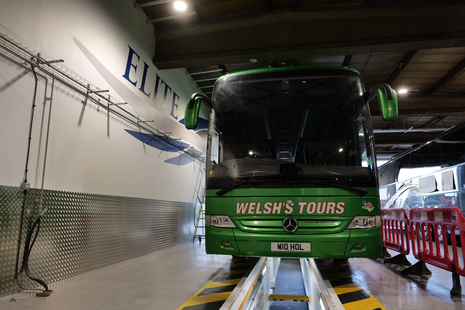 Green tour bus labeled 'Welsh's Tours' parked indoors next to a white wall with the word 'ELITE' and a blue arrow. Red barriers are set up beside the bus.